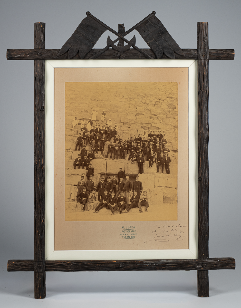 Fotografi inom glas och ram. Motiv: Svenska militärer och civila vid en pyramid i Kairo. Från korvetten Freja år 1890.
Ramen snidad med på översidan ett ankare med korslagda flaggor. Svartmålad.
Text: Till U. OK. Svensson. Minne från Cairo af (namnet oläsligt) 12.1. 90. G. DOGEA Photographe DE S.A. LE KHEDIVE PYRAMIDES. 1890.
Neg.nr 4076.