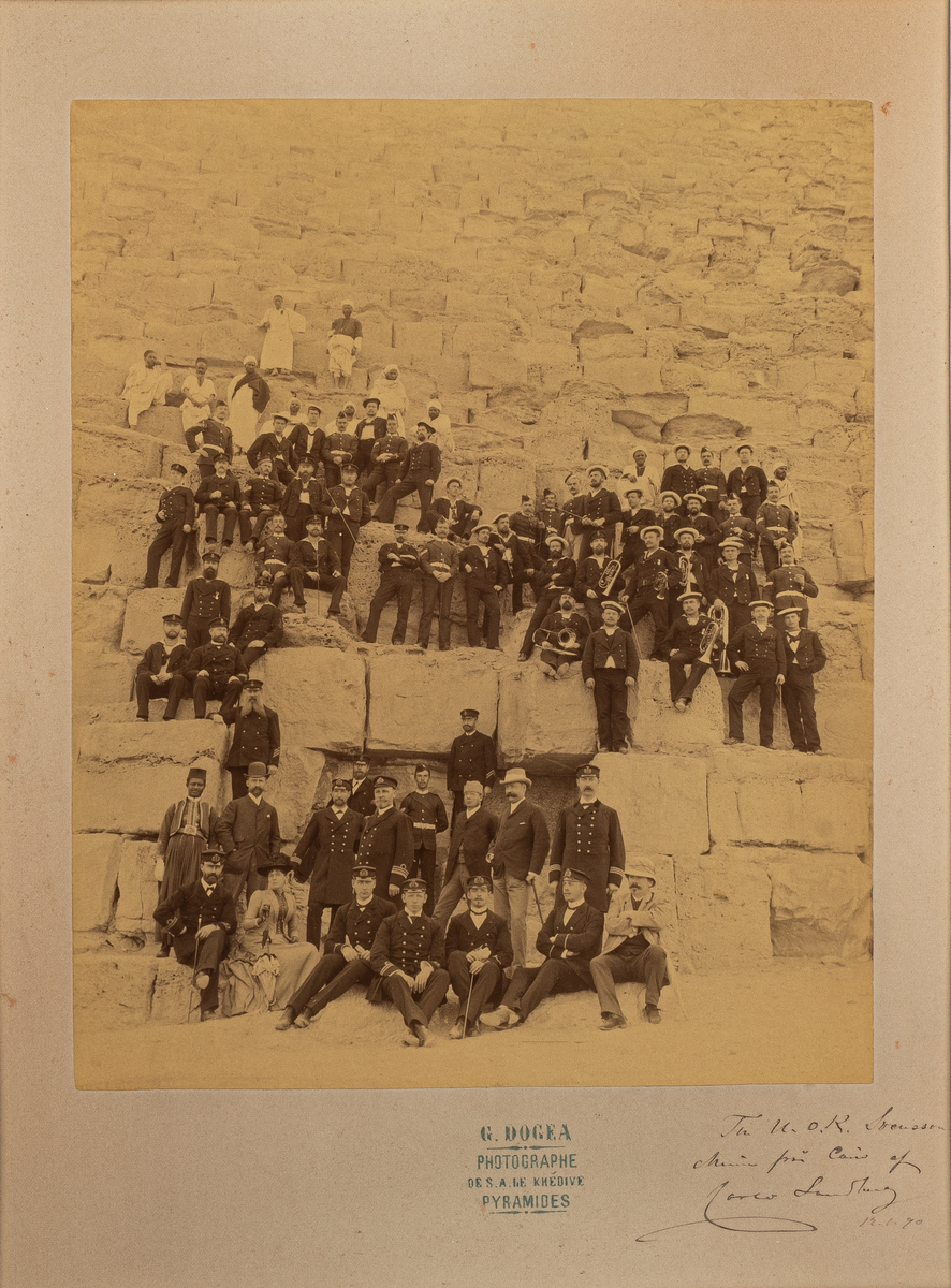Fotografi inom glas och ram. Motiv: Svenska militärer och civila vid en pyramid i Kairo. Från korvetten Freja år 1890.
Ramen snidad med på översidan ett ankare med korslagda flaggor. Svartmålad.
Text: Till U. OK. Svensson. Minne från Cairo af (namnet oläsligt) 12.1. 90. G. DOGEA Photographe DE S.A. LE KHEDIVE PYRAMIDES. 1890.
Neg.nr 4076.
