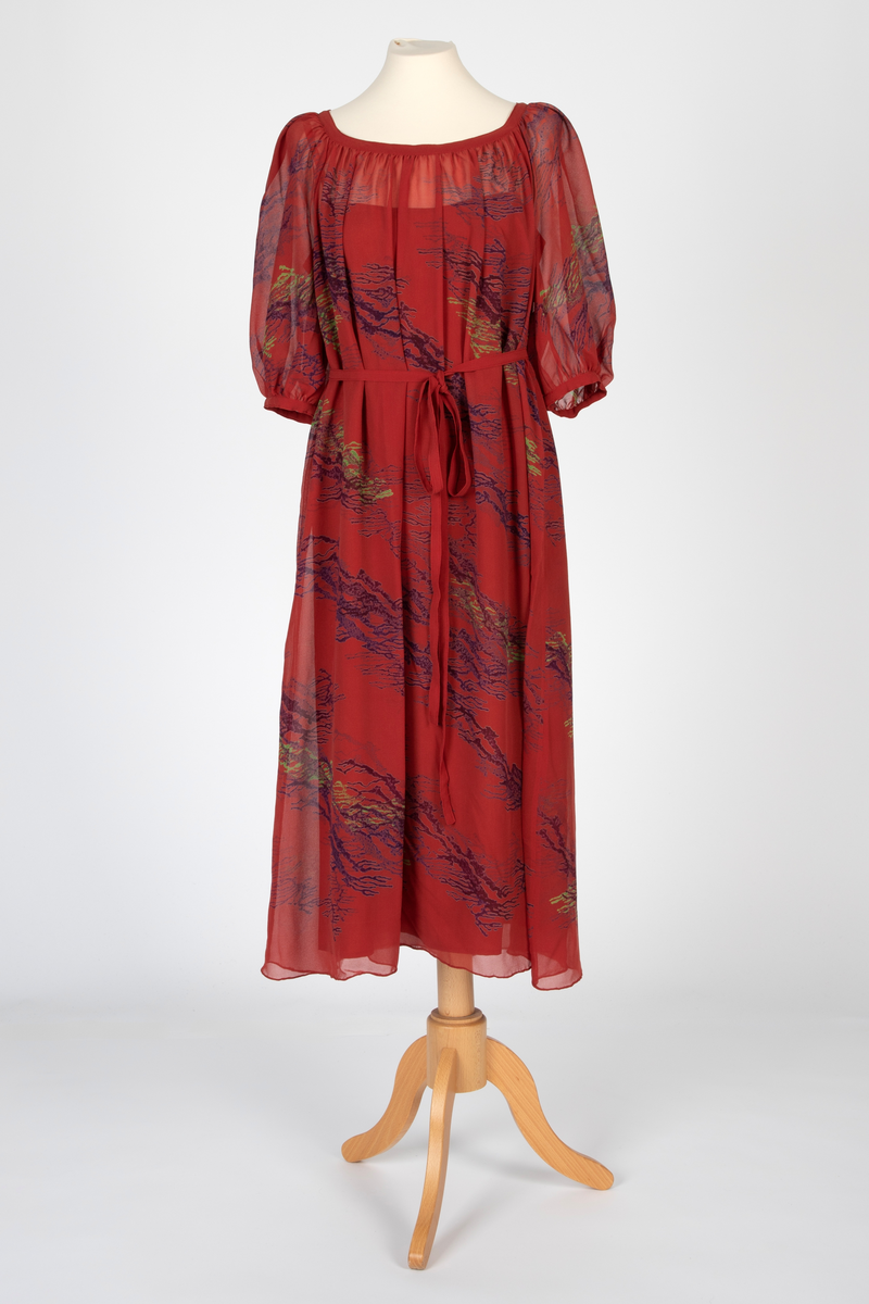 Kjole i natursilke, rødbrun bunnfarge med blokktrykkmønster i blåfiolett og grønt. Kjolen er helskåret, utskrådd og har halvlange, posete raglanermer. Båtformet utringing med smal linning. Herfra faller stoffet lett rynket til midt på leggen med rullefall nederst. Smalt belte som knyttes i livet. Underkjole av rødbrun taft med søm i hver side, utskrådd. Rett avskåret øverst og med smale, lysere bånd over skuldrene.