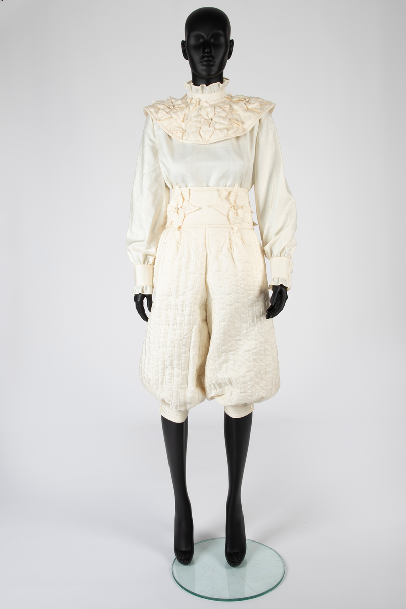 Ensemble i hvit kinesisk natursilke bestående av bluse, knikkers, løst belte og løs krage. Bukser, belte og krage er vattert, Mellomlegget i vatteringen er akrylvatt. Buksene er fôret med hvit syntetisk silke (japonette). Løsthengde bluse med hel rygg, sømmer i sidene og avrundet nederkant med splitter. Forside med bærestykker. Øverst knapping med perlemorsknapper og knapphull, nederst sammensydd. Oppstående halslinning med rysjekant. Lange ermer med knappet mansjett og rysjekant. Vatterte knikkers, fôret, med langsgående bisestikninger med cirka 2,5cm avstand. Åpning bak med glidelås og smal knappet livlinning. Bredere knappet knelinning. Stikklommer i sidene. Rektangulært, bredt belte, vattert og med stikninger langs kanten, knyttes ved to par sydde bånd av stoffet. Fire store blomster av stoffet, vatterte, er festet på beltet. Nesten sirkelrund krage med åpning foran og knyttebånd ved halsen. Vattert og med stikninger langs kantene. Fem store blomster med blad, samme som på beltet, er festet på kragen.