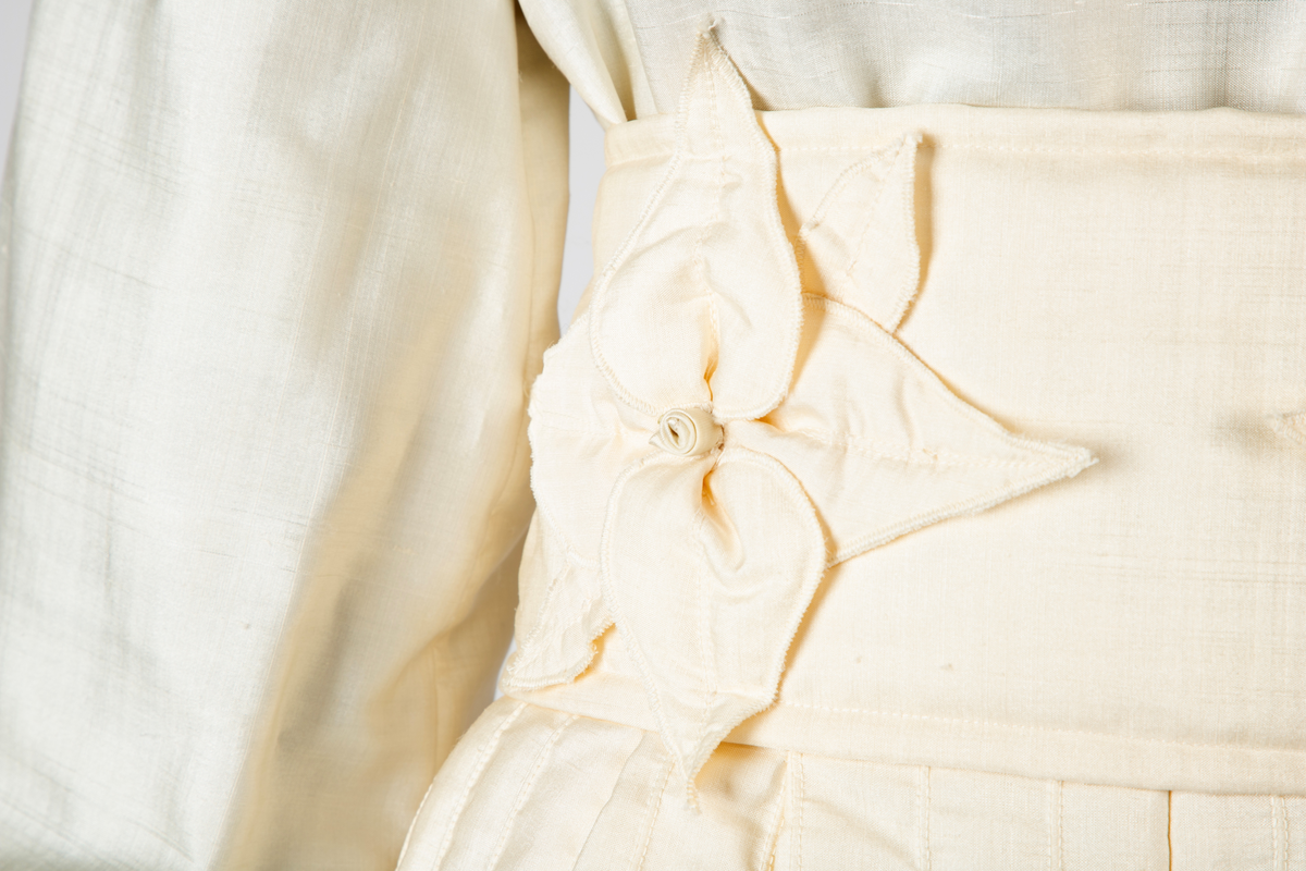 Ensemble i hvit kinesisk natursilke bestående av bluse, knikkers, løst belte og løs krage. Bukser, belte og krage er vattert, Mellomlegget i vatteringen er akrylvatt. Buksene er fôret med hvit syntetisk silke (japonette). Løsthengde bluse med hel rygg, sømmer i sidene og avrundet nederkant med splitter. Forside med bærestykker. Øverst knapping med perlemorsknapper og knapphull, nederst sammensydd. Oppstående halslinning med rysjekant. Lange ermer med knappet mansjett og rysjekant. Vatterte knikkers, fôret, med langsgående bisestikninger med cirka 2,5cm avstand. Åpning bak med glidelås og smal knappet livlinning. Bredere knappet knelinning. Stikklommer i sidene. Rektangulært, bredt belte, vattert og med stikninger langs kanten, knyttes ved to par sydde bånd av stoffet. Fire store blomster av stoffet, vatterte, er festet på beltet. Nesten sirkelrund krage med åpning foran og knyttebånd ved halsen. Vattert og med stikninger langs kantene. Fem store blomster med blad, samme som på beltet, er festet på kragen.