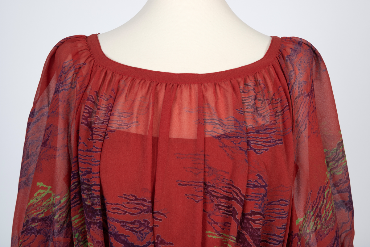 Kjole i natursilke, rødbrun bunnfarge med blokktrykkmønster i blåfiolett og grønt. Kjolen er helskåret, utskrådd og har halvlange, posete raglanermer. Båtformet utringing med smal linning. Herfra faller stoffet lett rynket til midt på leggen med rullefall nederst. Smalt belte som knyttes i livet. Underkjole av rødbrun taft med søm i hver side, utskrådd. Rett avskåret øverst og med smale, lysere bånd over skuldrene.