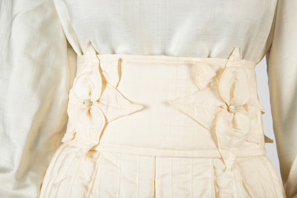 Ensemble i hvit kinesisk natursilke bestående av bluse, knikkers, løst belte og løs krage. Bukser, belte og krage er vattert, Mellomlegget i vatteringen er akrylvatt. Buksene er fôret med hvit syntetisk silke (japonette). Løsthengde bluse med hel rygg, sømmer i sidene og avrundet nederkant med splitter. Forside med bærestykker. Øverst knapping med perlemorsknapper og knapphull, nederst sammensydd. Oppstående halslinning med rysjekant. Lange ermer med knappet mansjett og rysjekant. Vatterte knikkers, fôret, med langsgående bisestikninger med cirka 2,5cm avstand. Åpning bak med glidelås og smal knappet livlinning. Bredere knappet knelinning. Stikklommer i sidene. Rektangulært, bredt belte, vattert og med stikninger langs kanten, knyttes ved to par sydde bånd av stoffet. Fire store blomster av stoffet, vatterte, er festet på beltet. Nesten sirkelrund krage med åpning foran og knyttebånd ved halsen. Vattert og med stikninger langs kantene. Fem store blomster med blad, samme som på beltet, er festet på kragen.