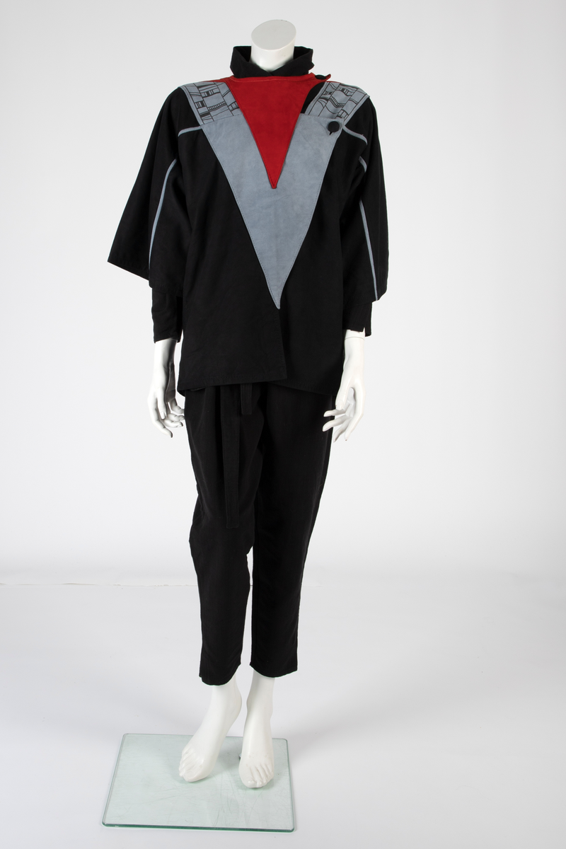 Kvinnedrakt bestående av jakke, overall og belte. Jakken er av svart semsket skinn, åpen foran og asymmetrisk knappet med en knapp på venstre skulder og en knapp i brysthøyde. Jakken har kimonolignende ermer med åpning i armhulene. Foran og bak, over skuldrene, delvis påsydd, delvis løsthengende grått skinn med påtrykt geometrisk mønster i svart. Det grå stykket går ned i en løsthengende spiss bak. Oppå skuldrene rødt semsket skinn. Over det grå stykket et lignende, men mindre stykke i rødt semsket skinn og bak et enda mindre stykke i svart semsket skinn, alle med spiss trekantform. Jakken er helfôret med syntetisk silke. Overallen er vid og av kraftig, svart bomullsstoff i ripsartet vevning. Overallen er åpen foran med skjult knapping. Buksebenene er vide øverst og smalner nedover med en liten splitt. Ermene er likedan. Påsydd krage med spisser. Til overallen hører et langt knyttebelte i samme stoff. Beltet er bredt på midten og smalner mot endene.