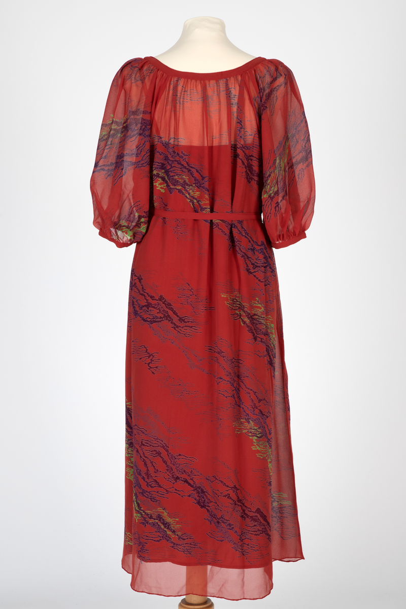 Kjole i natursilke, rødbrun bunnfarge med blokktrykkmønster i blåfiolett og grønt. Kjolen er helskåret, utskrådd og har halvlange, posete raglanermer. Båtformet utringing med smal linning. Herfra faller stoffet lett rynket til midt på leggen med rullefall nederst. Smalt belte som knyttes i livet. Underkjole av rødbrun taft med søm i hver side, utskrådd. Rett avskåret øverst og med smale, lysere bånd over skuldrene.