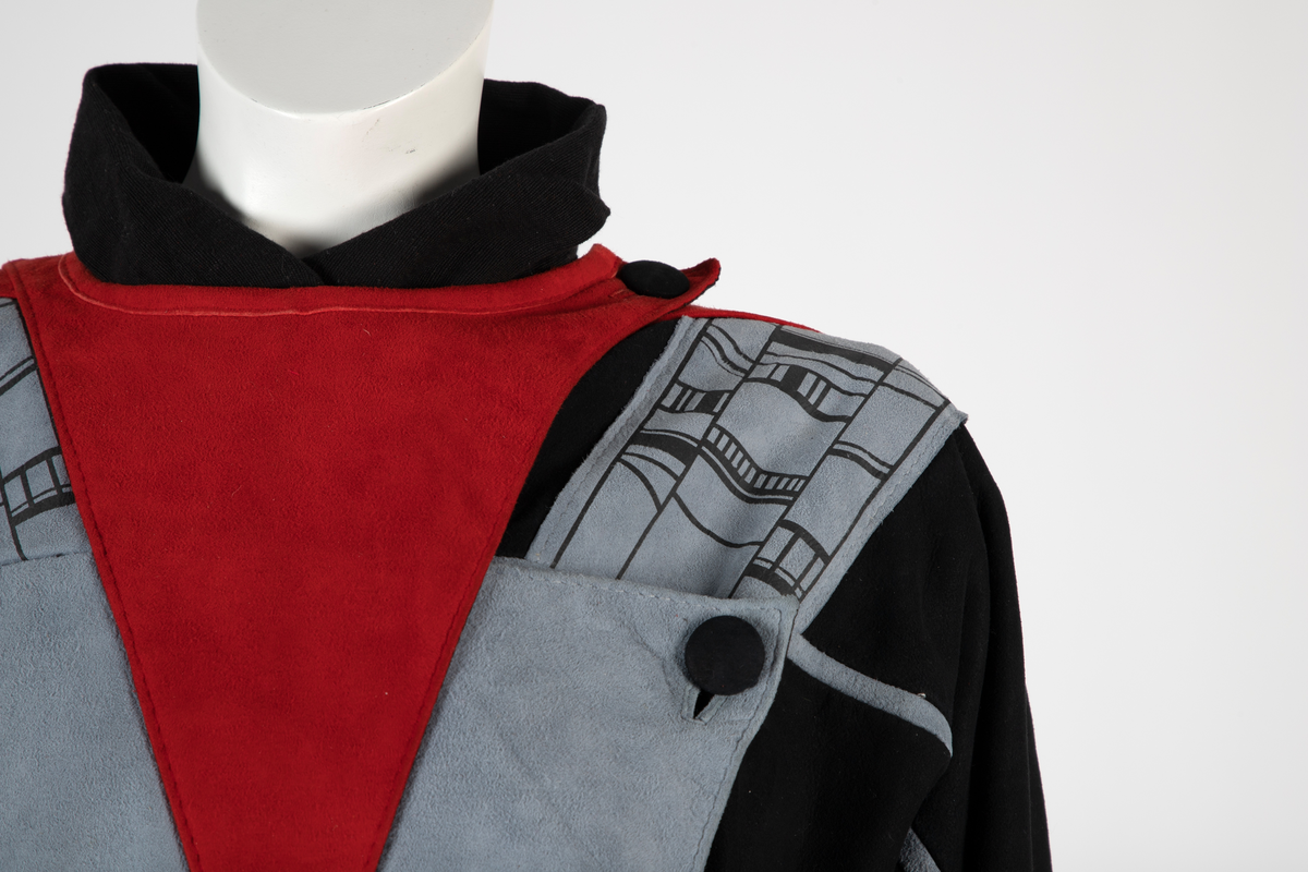 Kvinnedrakt bestående av jakke, overall og belte. Jakken er av svart semsket skinn, åpen foran og asymmetrisk knappet med en knapp på venstre skulder og en knapp i brysthøyde. Jakken har kimonolignende ermer med åpning i armhulene. Foran og bak, over skuldrene, delvis påsydd, delvis løsthengende grått skinn med påtrykt geometrisk mønster i svart. Det grå stykket går ned i en løsthengende spiss bak. Oppå skuldrene rødt semsket skinn. Over det grå stykket et lignende, men mindre stykke i rødt semsket skinn og bak et enda mindre stykke i svart semsket skinn, alle med spiss trekantform. Jakken er helfôret med syntetisk silke. Overallen er vid og av kraftig, svart bomullsstoff i ripsartet vevning. Overallen er åpen foran med skjult knapping. Buksebenene er vide øverst og smalner nedover med en liten splitt. Ermene er likedan. Påsydd krage med spisser. Til overallen hører et langt knyttebelte i samme stoff. Beltet er bredt på midten og smalner mot endene.