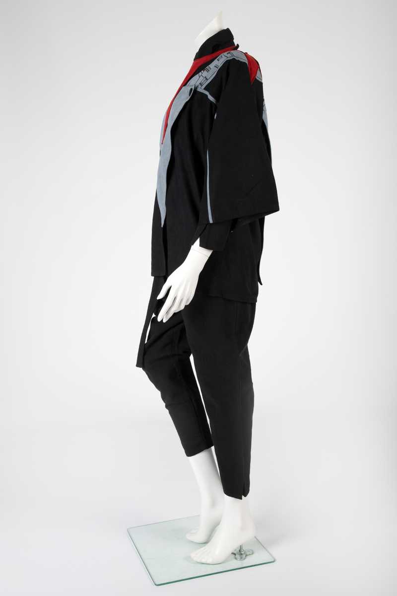 Kvinnedrakt bestående av jakke, overall og belte. Jakken er av svart semsket skinn, åpen foran og asymmetrisk knappet med en knapp på venstre skulder og en knapp i brysthøyde. Jakken har kimonolignende ermer med åpning i armhulene. Foran og bak, over skuldrene, delvis påsydd, delvis løsthengende grått skinn med påtrykt geometrisk mønster i svart. Det grå stykket går ned i en løsthengende spiss bak. Oppå skuldrene rødt semsket skinn. Over det grå stykket et lignende, men mindre stykke i rødt semsket skinn og bak et enda mindre stykke i svart semsket skinn, alle med spiss trekantform. Jakken er helfôret med syntetisk silke. Overallen er vid og av kraftig, svart bomullsstoff i ripsartet vevning. Overallen er åpen foran med skjult knapping. Buksebenene er vide øverst og smalner nedover med en liten splitt. Ermene er likedan. Påsydd krage med spisser. Til overallen hører et langt knyttebelte i samme stoff. Beltet er bredt på midten og smalner mot endene.