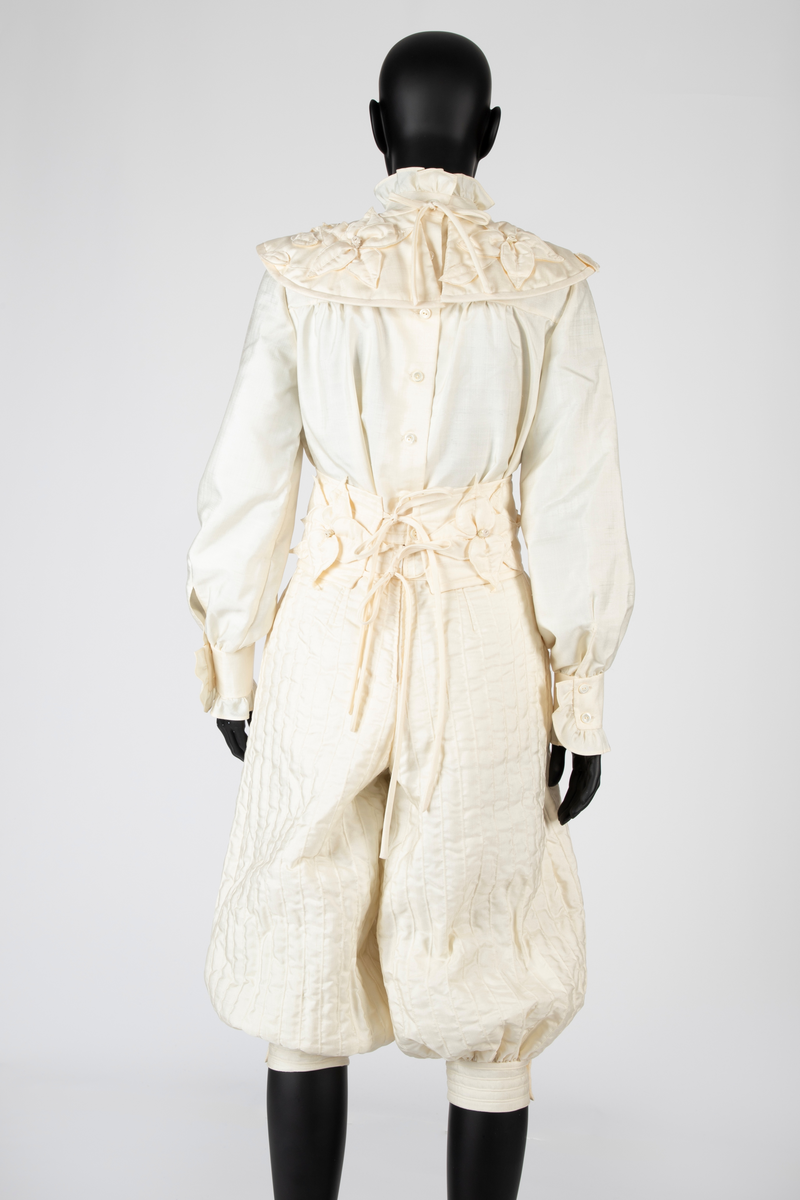 Ensemble i hvit kinesisk natursilke bestående av bluse, knikkers, løst belte og løs krage. Bukser, belte og krage er vattert, Mellomlegget i vatteringen er akrylvatt. Buksene er fôret med hvit syntetisk silke (japonette). Løsthengde bluse med hel rygg, sømmer i sidene og avrundet nederkant med splitter. Forside med bærestykker. Øverst knapping med perlemorsknapper og knapphull, nederst sammensydd. Oppstående halslinning med rysjekant. Lange ermer med knappet mansjett og rysjekant. Vatterte knikkers, fôret, med langsgående bisestikninger med cirka 2,5cm avstand. Åpning bak med glidelås og smal knappet livlinning. Bredere knappet knelinning. Stikklommer i sidene. Rektangulært, bredt belte, vattert og med stikninger langs kanten, knyttes ved to par sydde bånd av stoffet. Fire store blomster av stoffet, vatterte, er festet på beltet. Nesten sirkelrund krage med åpning foran og knyttebånd ved halsen. Vattert og med stikninger langs kantene. Fem store blomster med blad, samme som på beltet, er festet på kragen.