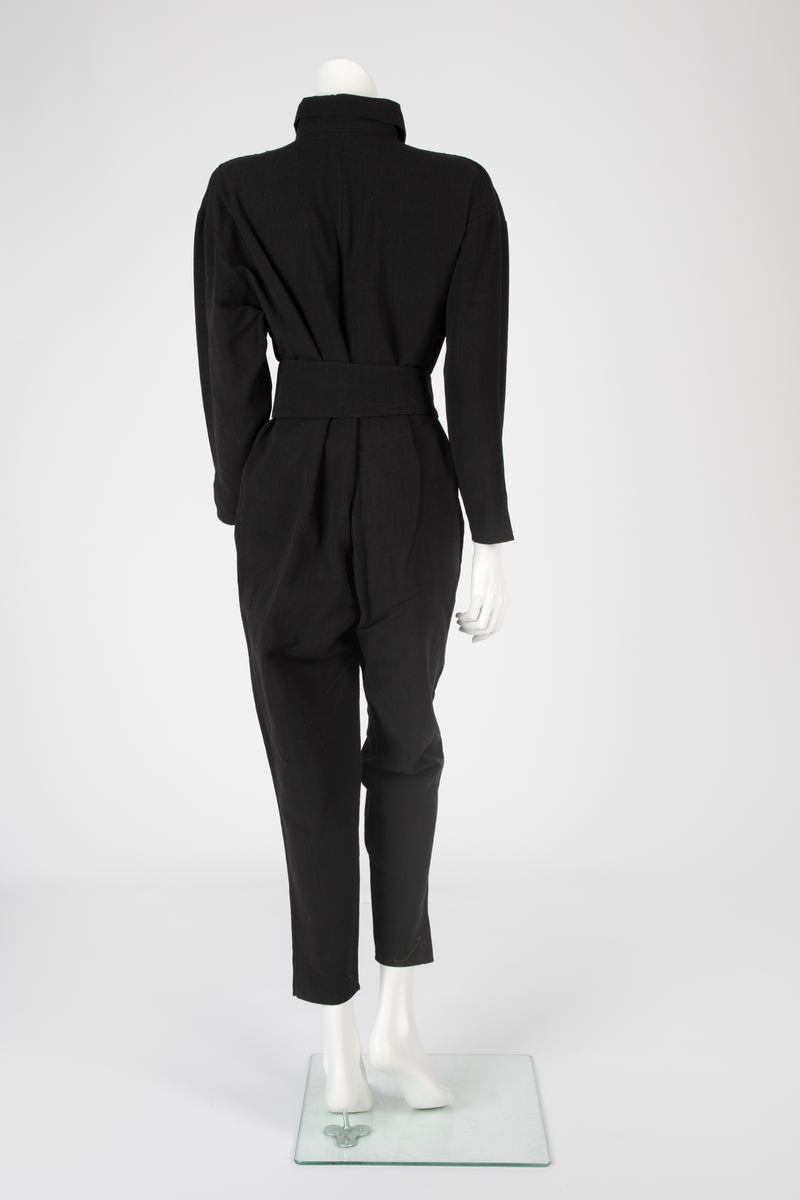 Kvinnedrakt bestående av jakke, overall og belte. Jakken er av svart semsket skinn, åpen foran og asymmetrisk knappet med en knapp på venstre skulder og en knapp i brysthøyde. Jakken har kimonolignende ermer med åpning i armhulene. Foran og bak, over skuldrene, delvis påsydd, delvis løsthengende grått skinn med påtrykt geometrisk mønster i svart. Det grå stykket går ned i en løsthengende spiss bak. Oppå skuldrene rødt semsket skinn. Over det grå stykket et lignende, men mindre stykke i rødt semsket skinn og bak et enda mindre stykke i svart semsket skinn, alle med spiss trekantform. Jakken er helfôret med syntetisk silke. Overallen er vid og av kraftig, svart bomullsstoff i ripsartet vevning. Overallen er åpen foran med skjult knapping. Buksebenene er vide øverst og smalner nedover med en liten splitt. Ermene er likedan. Påsydd krage med spisser. Til overallen hører et langt knyttebelte i samme stoff. Beltet er bredt på midten og smalner mot endene.