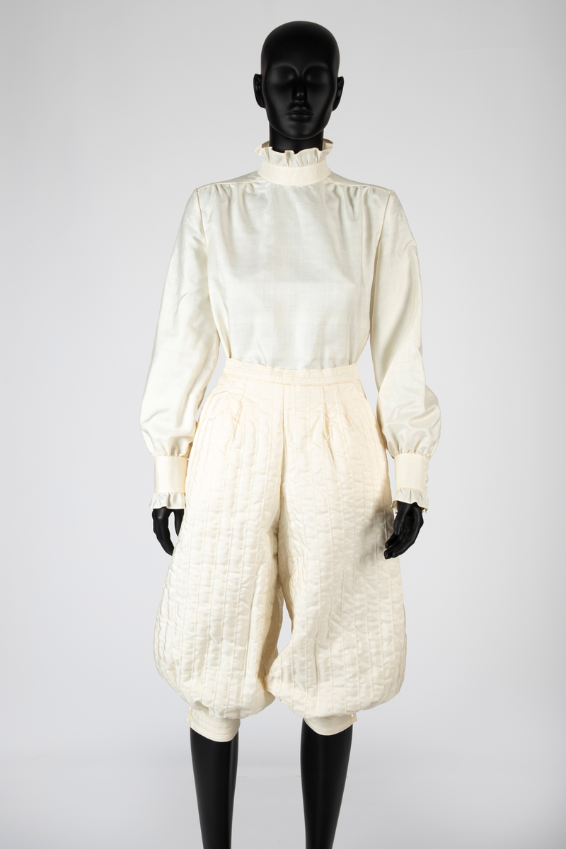 Ensemble i hvit kinesisk natursilke bestående av bluse, knikkers, løst belte og løs krage. Bukser, belte og krage er vattert, Mellomlegget i vatteringen er akrylvatt. Buksene er fôret med hvit syntetisk silke (japonette). Løsthengde bluse med hel rygg, sømmer i sidene og avrundet nederkant med splitter. Forside med bærestykker. Øverst knapping med perlemorsknapper og knapphull, nederst sammensydd. Oppstående halslinning med rysjekant. Lange ermer med knappet mansjett og rysjekant. Vatterte knikkers, fôret, med langsgående bisestikninger med cirka 2,5cm avstand. Åpning bak med glidelås og smal knappet livlinning. Bredere knappet knelinning. Stikklommer i sidene. Rektangulært, bredt belte, vattert og med stikninger langs kanten, knyttes ved to par sydde bånd av stoffet. Fire store blomster av stoffet, vatterte, er festet på beltet. Nesten sirkelrund krage med åpning foran og knyttebånd ved halsen. Vattert og med stikninger langs kantene. Fem store blomster med blad, samme som på beltet, er festet på kragen.