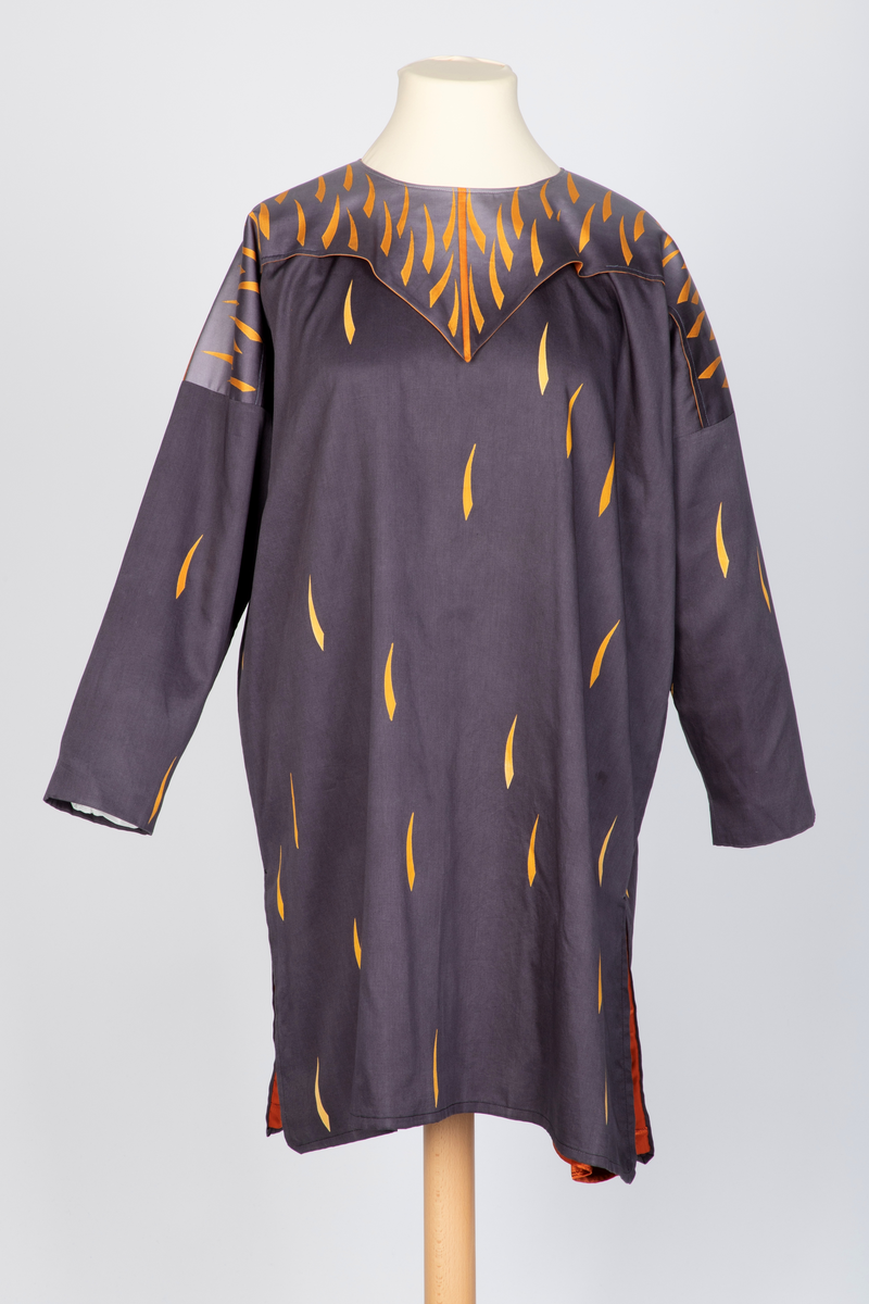 Ytterplagg i bomull med stofftrykk; mørk fiolettblått med gule og oransjefargede, lange, krummede spetter. Fôret med oransjefarget, syntetisk silke. Vid, kimonolignende fasong med søm i hver side og splitt nederst. Lange ermer og vid, rundet halsrigning. Plagget trekkes over hodet. Løst over skuldrene et cape- eller kragelignende stykke stoff som går ned i en spiss foran og i en dypere spiss bak, fastsydd til halsåpningen og til ermesømmene. Dette stykket går over i en lysere sjattering bunnfarge mot halsen og har et tettere spettemønster. Oransje fôr.