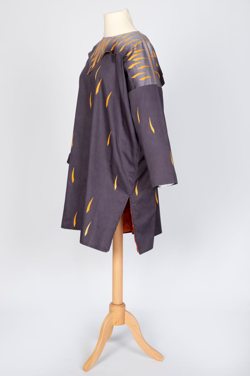 Ytterplagg i bomull med stofftrykk; mørk fiolettblått med gule og oransjefargede, lange, krummede spetter. Fôret med oransjefarget, syntetisk silke. Vid, kimonolignende fasong med søm i hver side og splitt nederst. Lange ermer og vid, rundet halsrigning. Plagget trekkes over hodet. Løst over skuldrene et cape- eller kragelignende stykke stoff som går ned i en spiss foran og i en dypere spiss bak, fastsydd til halsåpningen og til ermesømmene. Dette stykket går over i en lysere sjattering bunnfarge mot halsen og har et tettere spettemønster. Oransje fôr.
