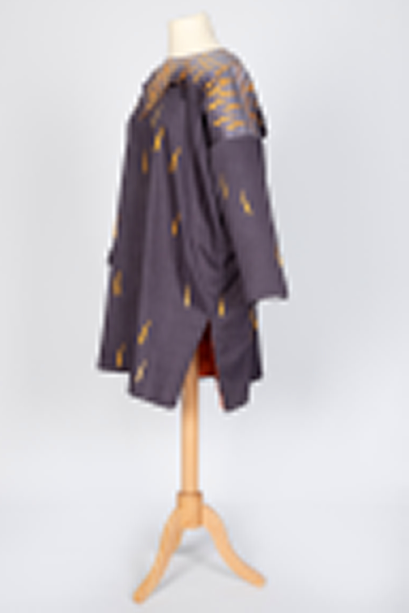 Ytterplagg i bomull med stofftrykk; mørk fiolettblått med gule og oransjefargede, lange, krummede spetter. Fôret med oransjefarget, syntetisk silke. Vid, kimonolignende fasong med søm i hver side og splitt nederst. Lange ermer og vid, rundet halsrigning. Plagget trekkes over hodet. Løst over skuldrene et cape- eller kragelignende stykke stoff som går ned i en spiss foran og i en dypere spiss bak, fastsydd til halsåpningen og til ermesømmene. Dette stykket går over i en lysere sjattering bunnfarge mot halsen og har et tettere spettemønster. Oransje fôr.