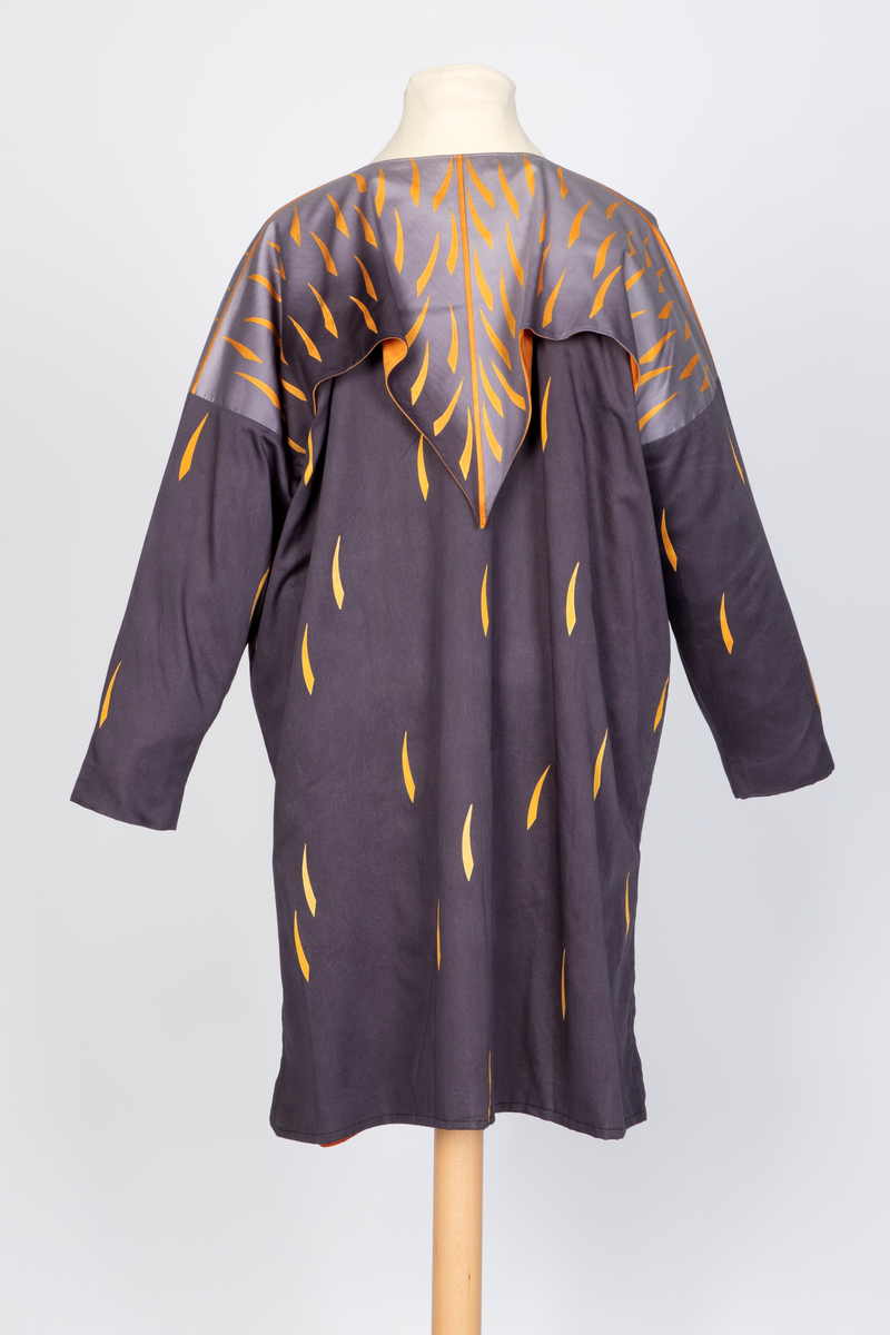 Ytterplagg i bomull med stofftrykk; mørk fiolettblått med gule og oransjefargede, lange, krummede spetter. Fôret med oransjefarget, syntetisk silke. Vid, kimonolignende fasong med søm i hver side og splitt nederst. Lange ermer og vid, rundet halsrigning. Plagget trekkes over hodet. Løst over skuldrene et cape- eller kragelignende stykke stoff som går ned i en spiss foran og i en dypere spiss bak, fastsydd til halsåpningen og til ermesømmene. Dette stykket går over i en lysere sjattering bunnfarge mot halsen og har et tettere spettemønster. Oransje fôr.