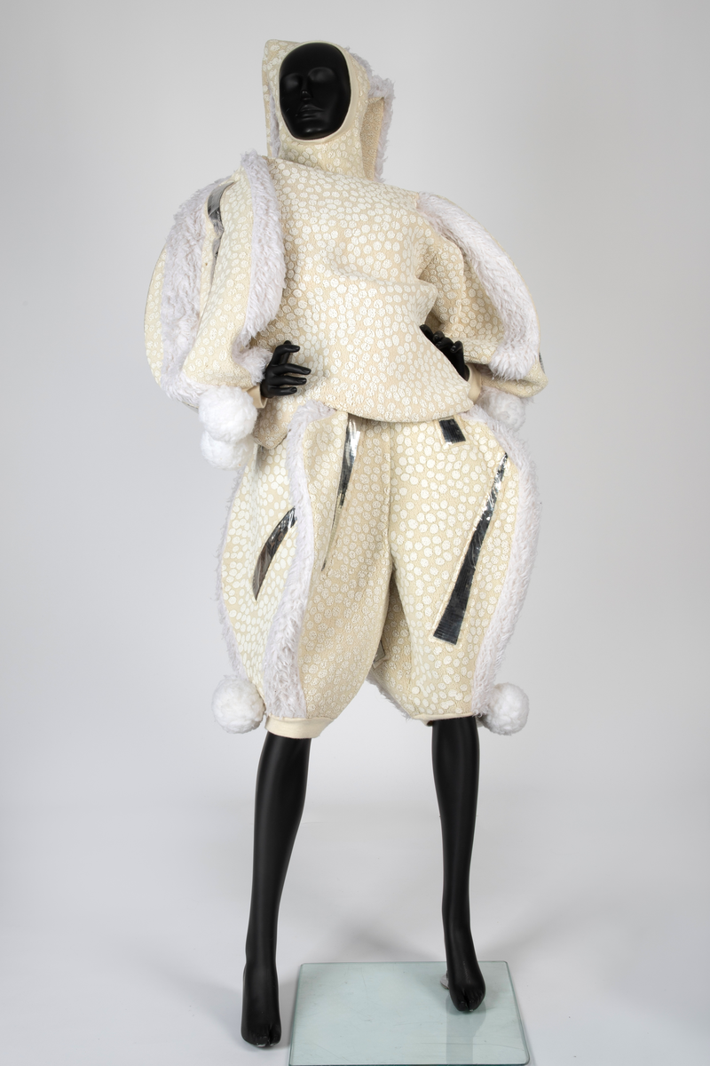 Overdel og selebukse i gulhvit, syntetisk fleece dekorert med plastspeil. Selebuksen har vide ben til knærne. Overdelen har vide ermer og går i ett med hetten som har to topper.