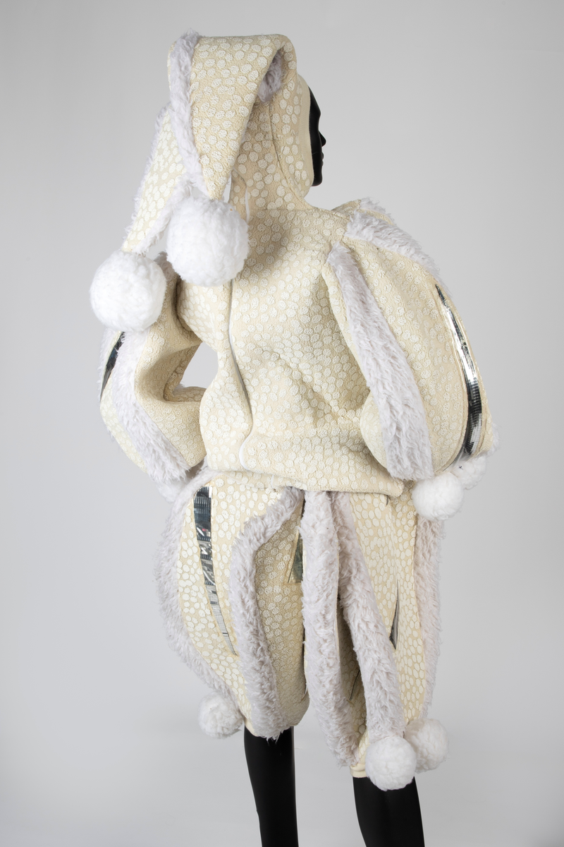 Overdel og selebukse i gulhvit, syntetisk fleece dekorert med plastspeil. Selebuksen har vide ben til knærne. Overdelen har vide ermer og går i ett med hetten som har to topper.