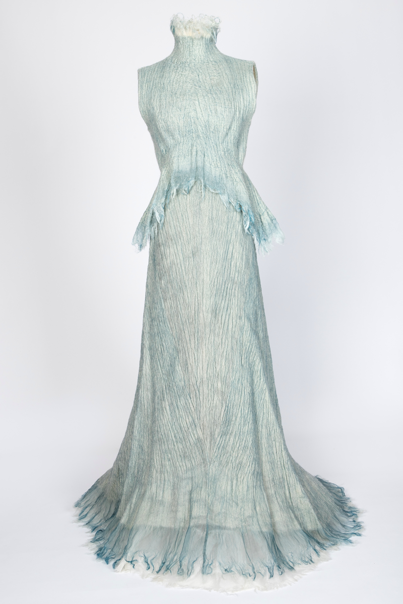 Kjole og overdel i tovet lys turkis-blå-grå ull og silkeorgansa med innslag av metalltråd. Benhvitt underskjørt av silkeorganza med metallstreng i nederkant.