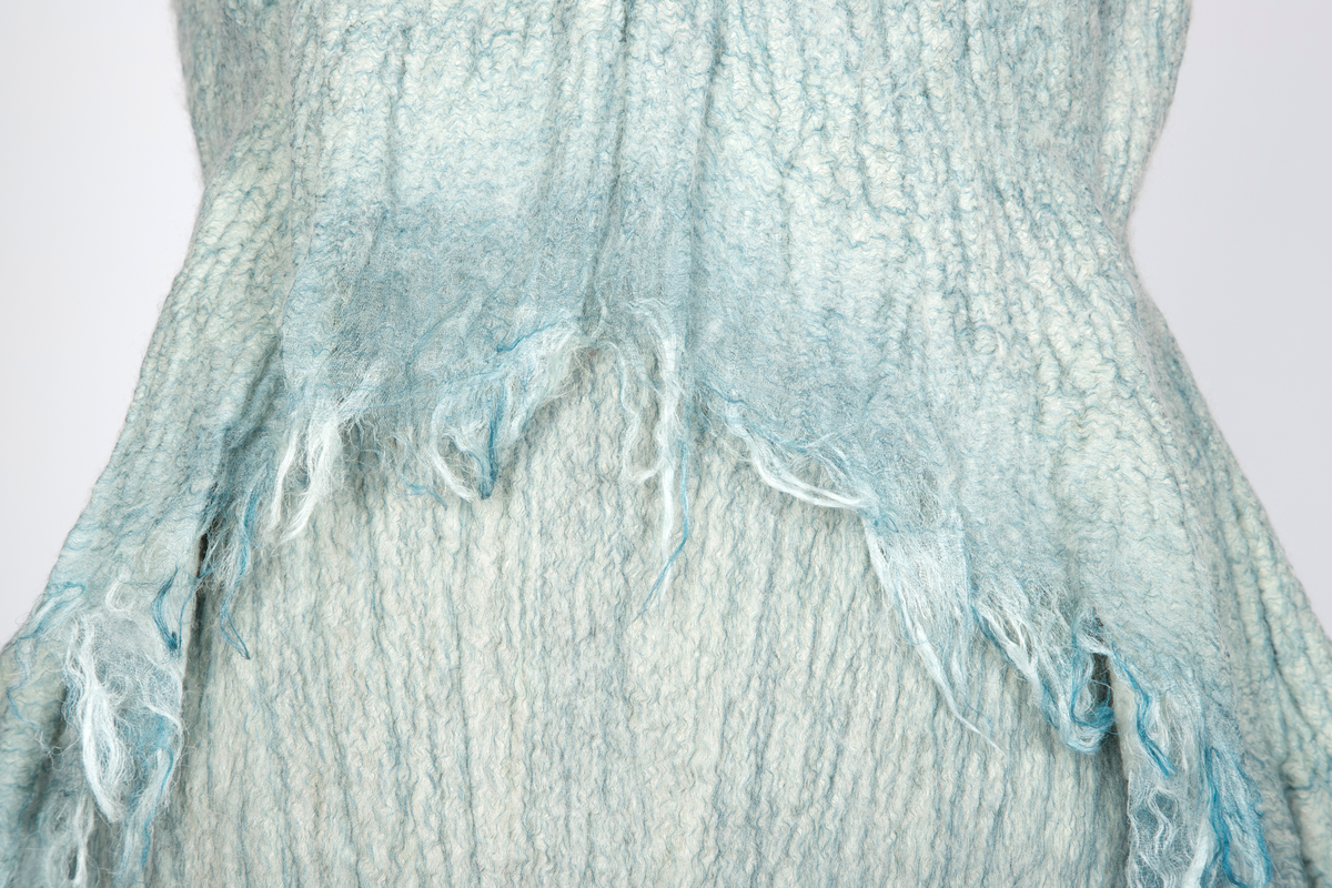 Kjole og overdel i tovet lys turkis-blå-grå ull og silkeorgansa med innslag av metalltråd. Benhvitt underskjørt av silkeorganza med metallstreng i nederkant.