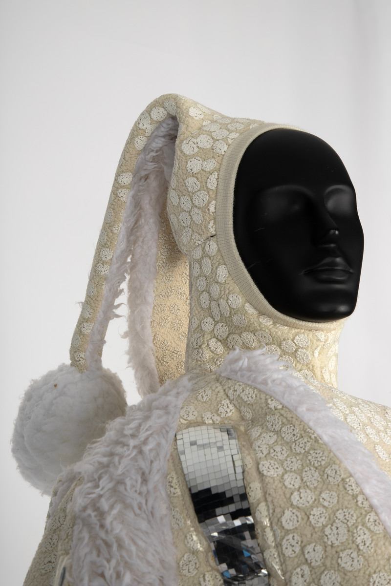 Overdel og selebukse i gulhvit, syntetisk fleece dekorert med plastspeil. Selebuksen har vide ben til knærne. Overdelen har vide ermer og går i ett med hetten som har to topper.