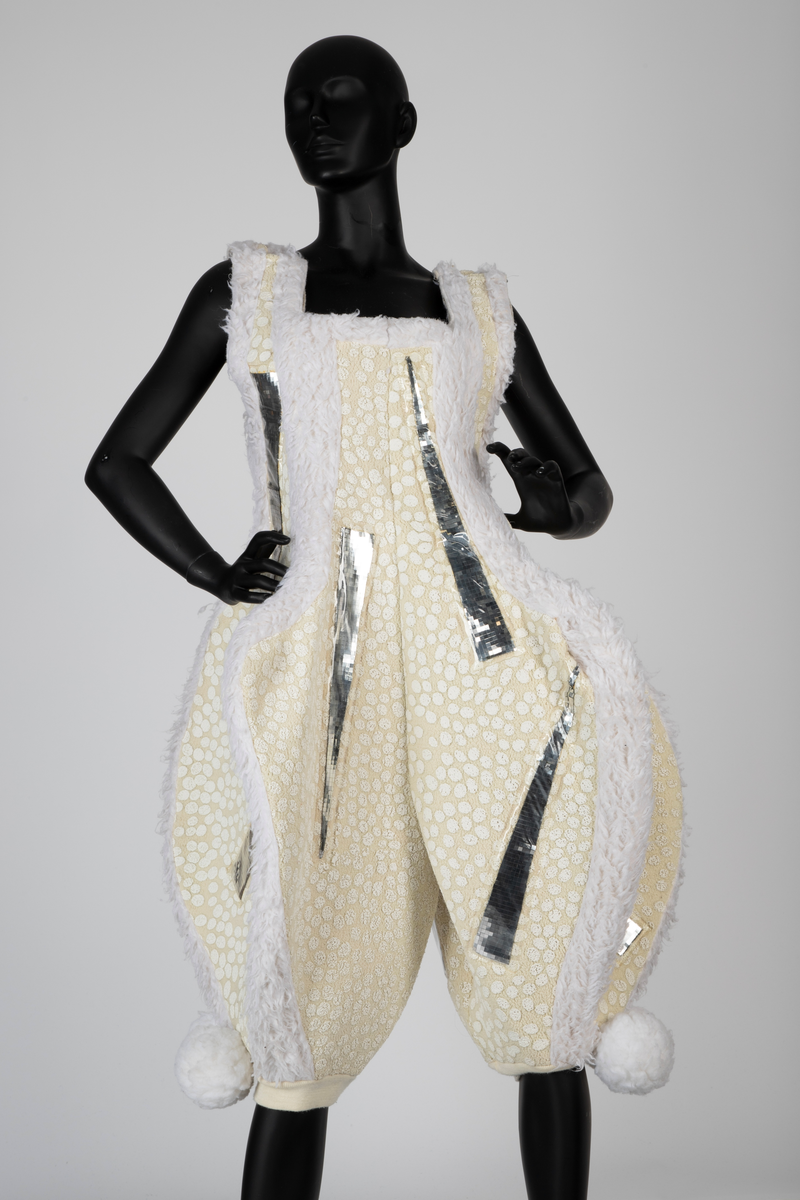 Overdel og selebukse i gulhvit, syntetisk fleece dekorert med plastspeil. Selebuksen har vide ben til knærne. Overdelen har vide ermer og går i ett med hetten som har to topper.