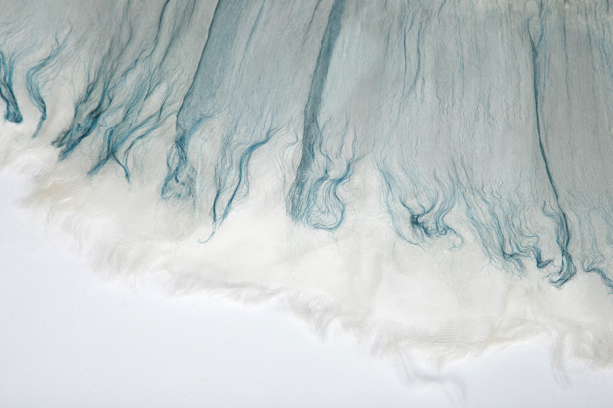 Kjole og overdel i tovet lys turkis-blå-grå ull og silkeorgansa med innslag av metalltråd. Benhvitt underskjørt av silkeorganza med metallstreng i nederkant.