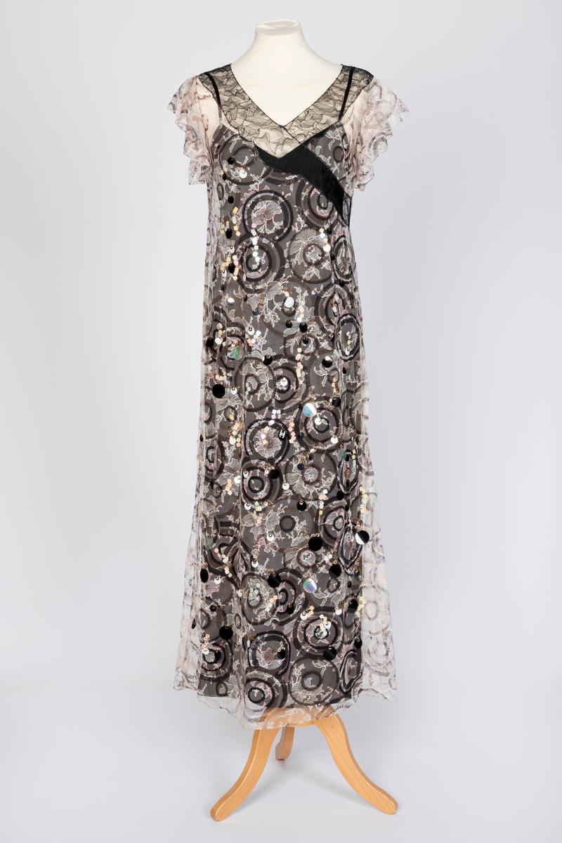 Fotsid kjole med tilhørende liten hatt. Kjolen består av en svart underkjole i silke med v-formet utringning og smale skulderstropper og en overkjole av gjennomsiktig, mønstertrykt tyllstoff med blonder. Mønsteret består av svarte, brune, beige, hvite og rosa sirkler. Rund utringning og korte, volangeformede ermer. Kjolen har påsydde paljetter og Chanels monogram i matchende farger. Tilhørende liten silkechiffonhatt i solhattfasong, påtrykt samme mønster som kjolen.