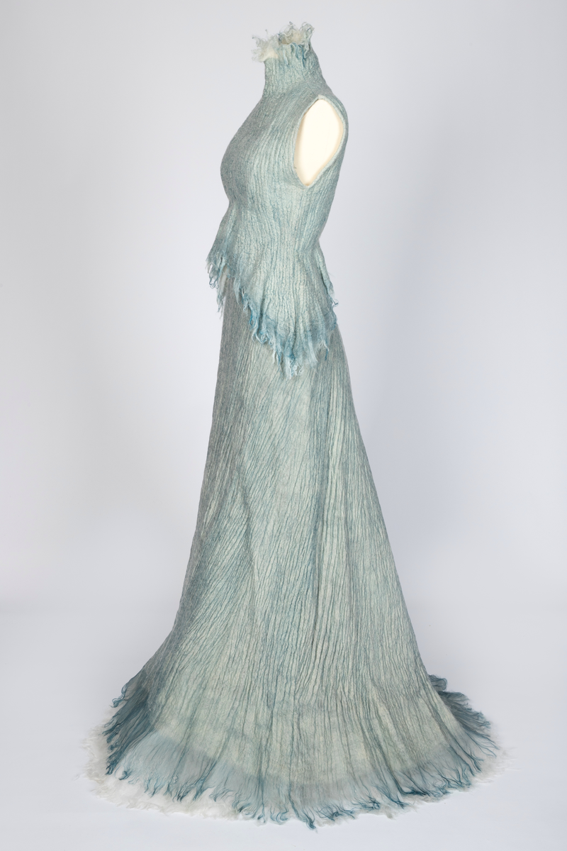 Kjole og overdel i tovet lys turkis-blå-grå ull og silkeorgansa med innslag av metalltråd. Benhvitt underskjørt av silkeorganza med metallstreng i nederkant.