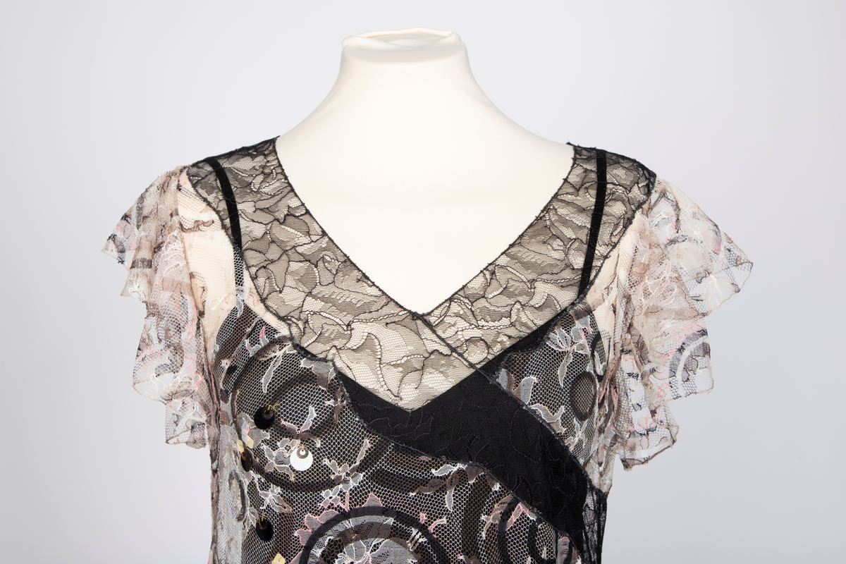 Fotsid kjole med tilhørende liten hatt. Kjolen består av en svart underkjole i silke med v-formet utringning og smale skulderstropper og en overkjole av gjennomsiktig, mønstertrykt tyllstoff med blonder. Mønsteret består av svarte, brune, beige, hvite og rosa sirkler. Rund utringning og korte, volangeformede ermer. Kjolen har påsydde paljetter og Chanels monogram i matchende farger. Tilhørende liten silkechiffonhatt i solhattfasong, påtrykt samme mønster som kjolen.