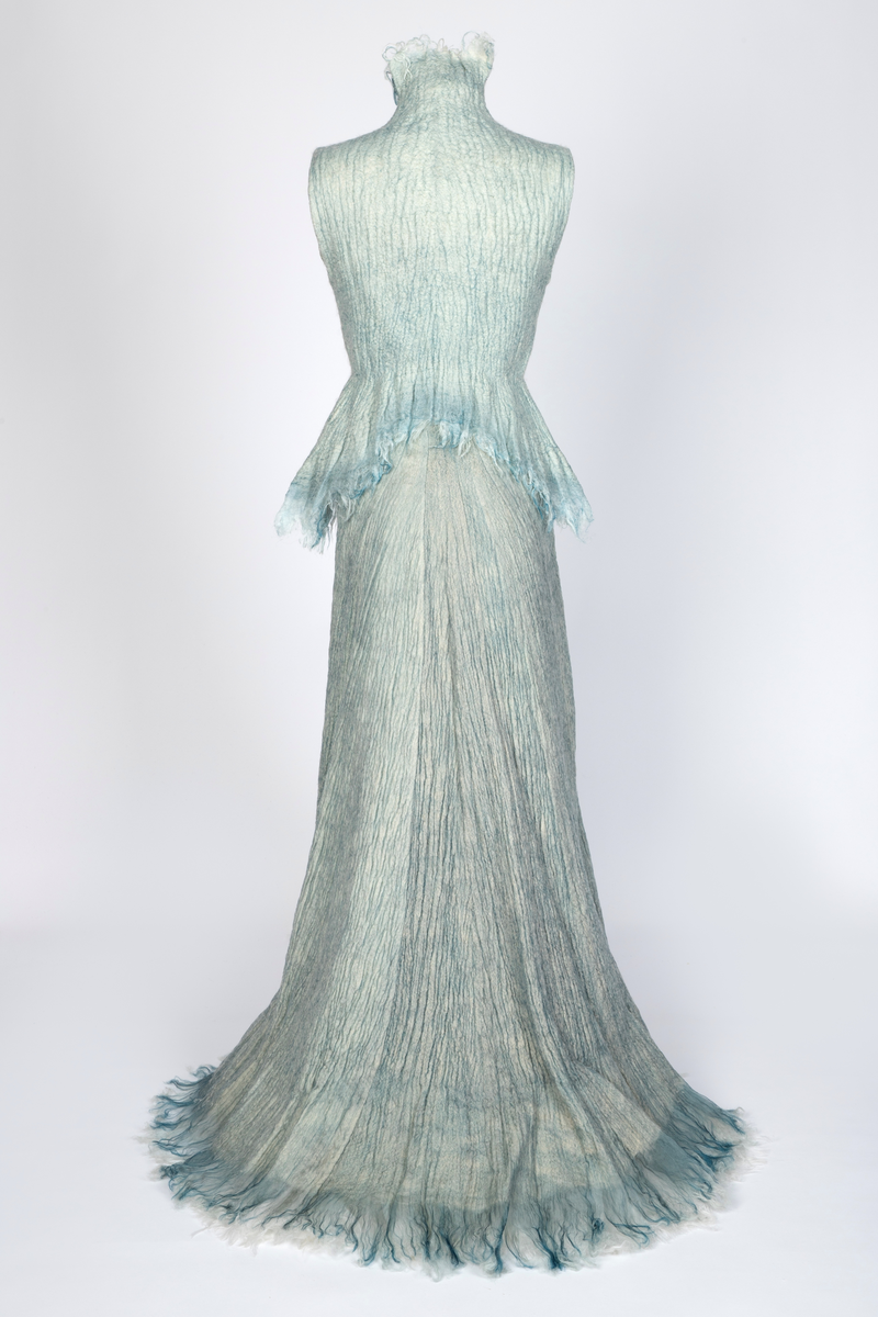 Kjole og overdel i tovet lys turkis-blå-grå ull og silkeorgansa med innslag av metalltråd. Benhvitt underskjørt av silkeorganza med metallstreng i nederkant.