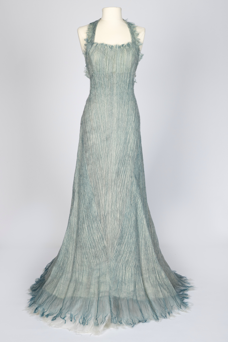 Kjole og overdel i tovet lys turkis-blå-grå ull og silkeorgansa med innslag av metalltråd. Benhvitt underskjørt av silkeorganza med metallstreng i nederkant.