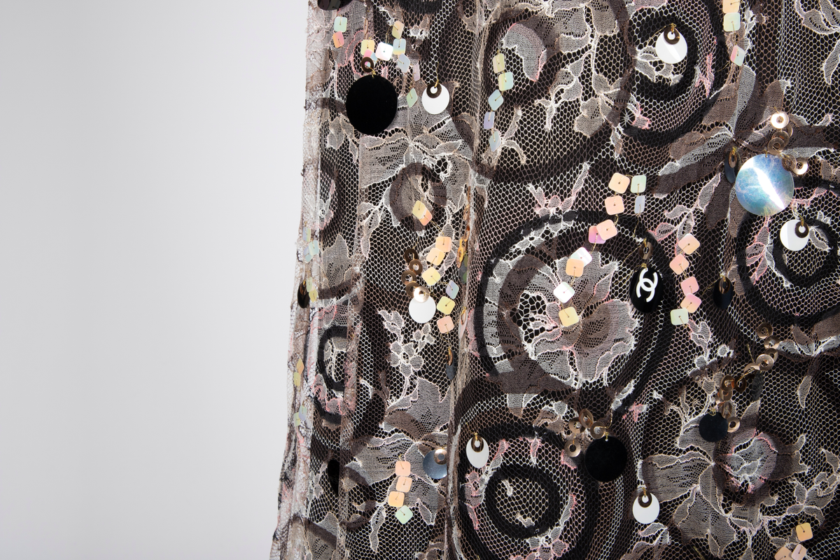 Fotsid kjole med tilhørende liten hatt. Kjolen består av en svart underkjole i silke med v-formet utringning og smale skulderstropper og en overkjole av gjennomsiktig, mønstertrykt tyllstoff med blonder. Mønsteret består av svarte, brune, beige, hvite og rosa sirkler. Rund utringning og korte, volangeformede ermer. Kjolen har påsydde paljetter og Chanels monogram i matchende farger. Tilhørende liten silkechiffonhatt i solhattfasong, påtrykt samme mønster som kjolen.