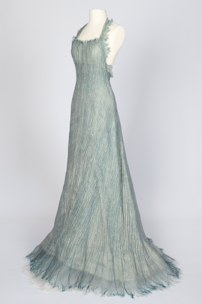 Kjole og overdel i tovet lys turkis-blå-grå ull og silkeorgansa med innslag av metalltråd. Benhvitt underskjørt av silkeorganza med metallstreng i nederkant.