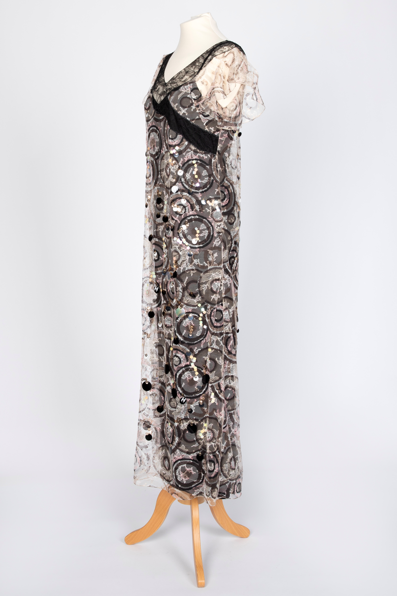 Fotsid kjole med tilhørende liten hatt. Kjolen består av en svart underkjole i silke med v-formet utringning og smale skulderstropper og en overkjole av gjennomsiktig, mønstertrykt tyllstoff med blonder. Mønsteret består av svarte, brune, beige, hvite og rosa sirkler. Rund utringning og korte, volangeformede ermer. Kjolen har påsydde paljetter og Chanels monogram i matchende farger. Tilhørende liten silkechiffonhatt i solhattfasong, påtrykt samme mønster som kjolen.