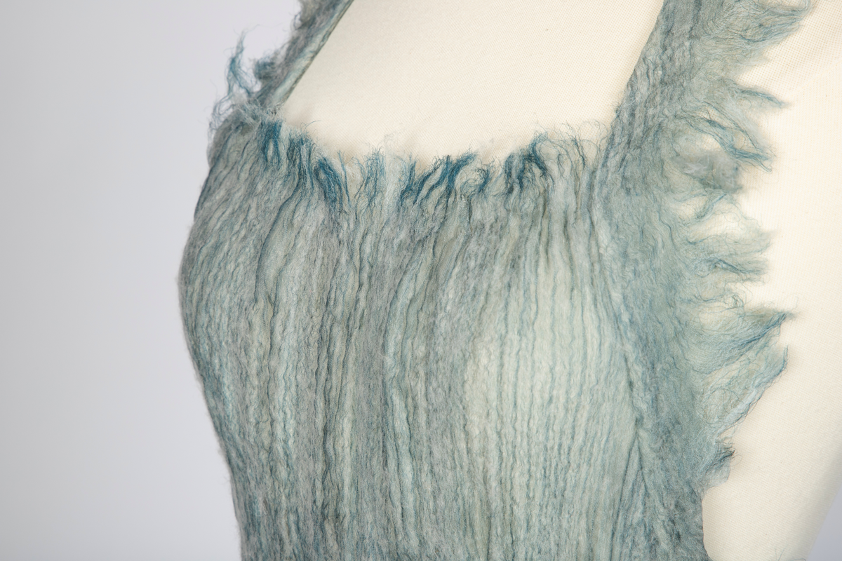 Kjole og overdel i tovet lys turkis-blå-grå ull og silkeorgansa med innslag av metalltråd. Benhvitt underskjørt av silkeorganza med metallstreng i nederkant.