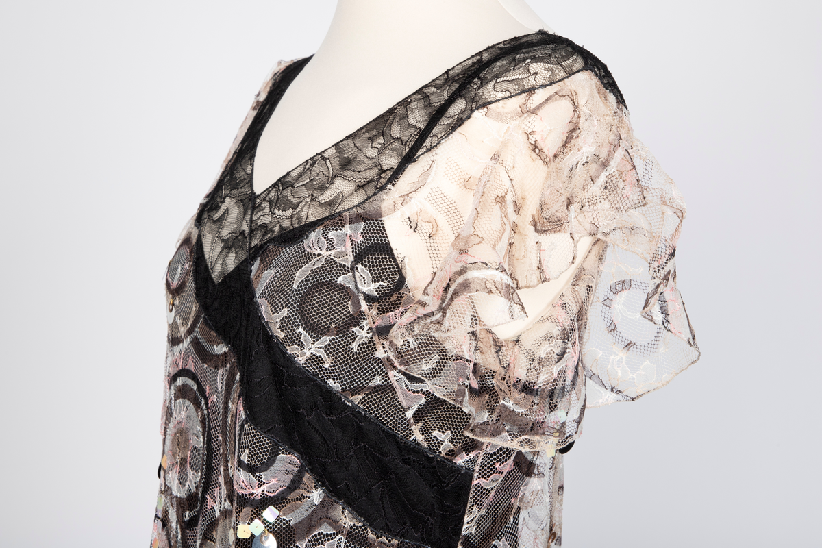 Fotsid kjole med tilhørende liten hatt. Kjolen består av en svart underkjole i silke med v-formet utringning og smale skulderstropper og en overkjole av gjennomsiktig, mønstertrykt tyllstoff med blonder. Mønsteret består av svarte, brune, beige, hvite og rosa sirkler. Rund utringning og korte, volangeformede ermer. Kjolen har påsydde paljetter og Chanels monogram i matchende farger. Tilhørende liten silkechiffonhatt i solhattfasong, påtrykt samme mønster som kjolen.