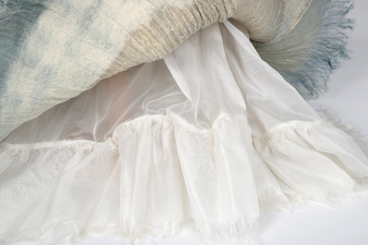 Kjole og overdel i tovet lys turkis-blå-grå ull og silkeorgansa med innslag av metalltråd. Benhvitt underskjørt av silkeorganza med metallstreng i nederkant.