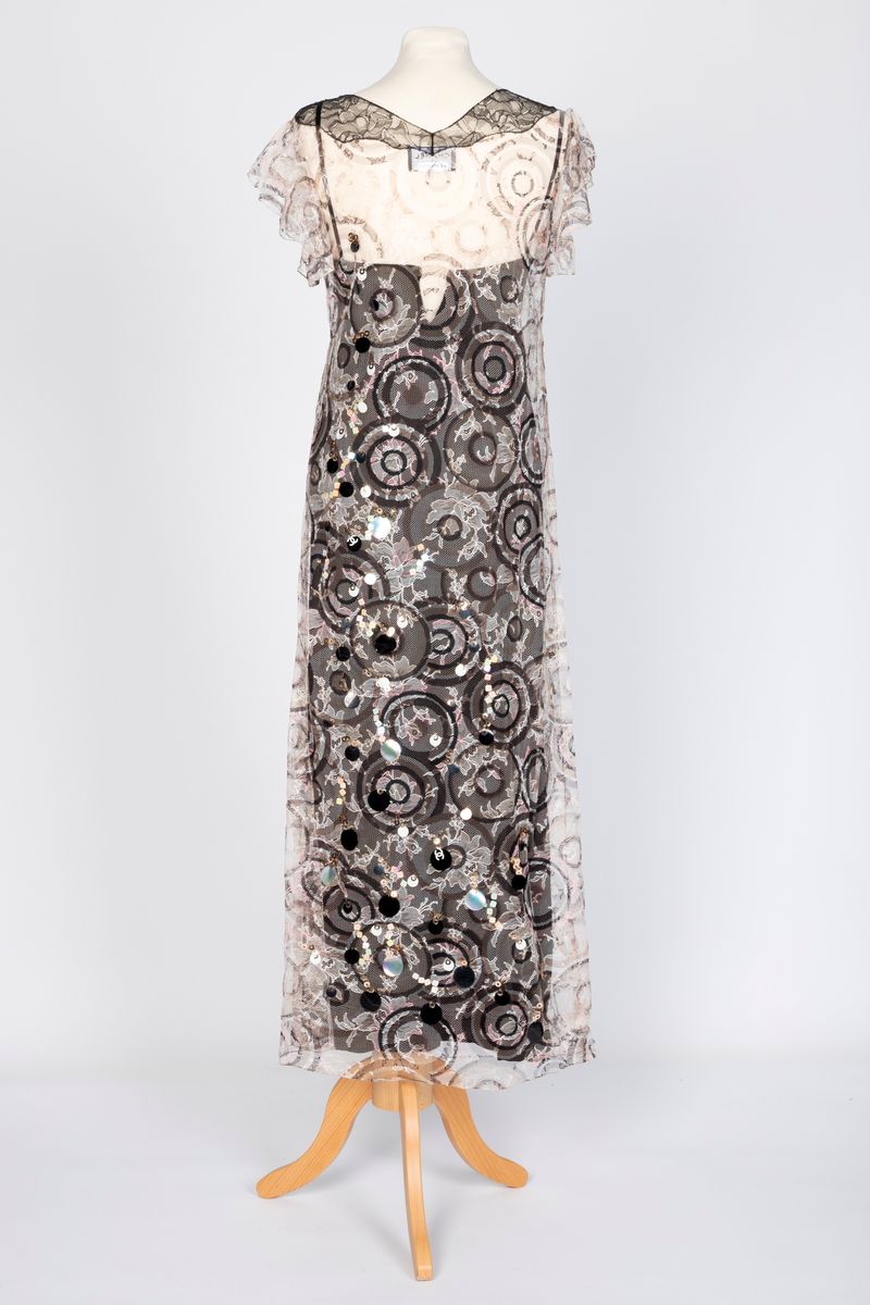 Fotsid kjole med tilhørende liten hatt. Kjolen består av en svart underkjole i silke med v-formet utringning og smale skulderstropper og en overkjole av gjennomsiktig, mønstertrykt tyllstoff med blonder. Mønsteret består av svarte, brune, beige, hvite og rosa sirkler. Rund utringning og korte, volangeformede ermer. Kjolen har påsydde paljetter og Chanels monogram i matchende farger. Tilhørende liten silkechiffonhatt i solhattfasong, påtrykt samme mønster som kjolen.