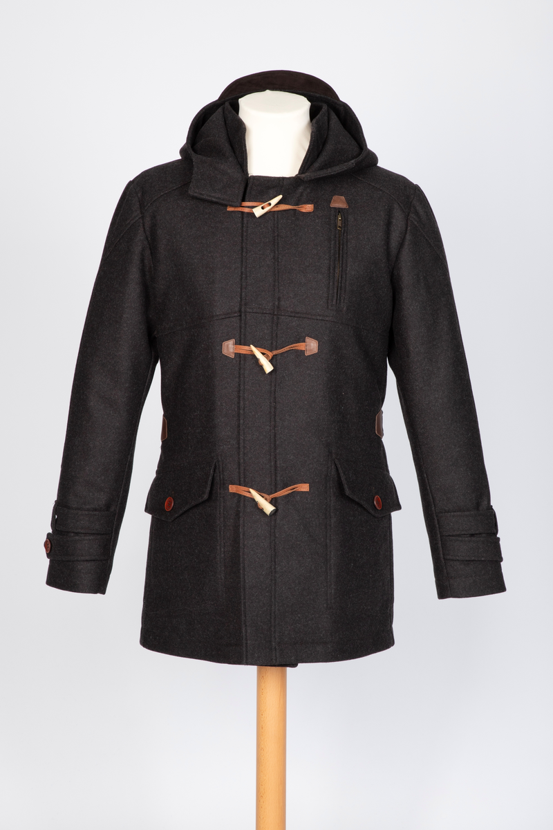 Dyffel med hette i brun ull. Bærestykke foran, på skuldrene og et bærestykke i skinnlignende materiale midt bak. Glidelås med klaff midt foran. Til lukking tre skinnsnorer med en beinlignende knapp. Store innfelte lommer med stor klaff med knapphull og gyllenbrun knapp. Søm midt bak med splitt, og formende panelsømmer. Belte festet rett foran sidesømmen i livet. Tosøms-ermer, dobbelklaff med knapp og knapphull og tre hemper på øverste klaffen. Sømmen på skulderens bærestykke fortsetter over til erme, parallelt nedover til den skrår inn tre centimeter før klaffene. Rødt, rutestukket, vattert fôr. Jakken er laget med nano-teknologi.