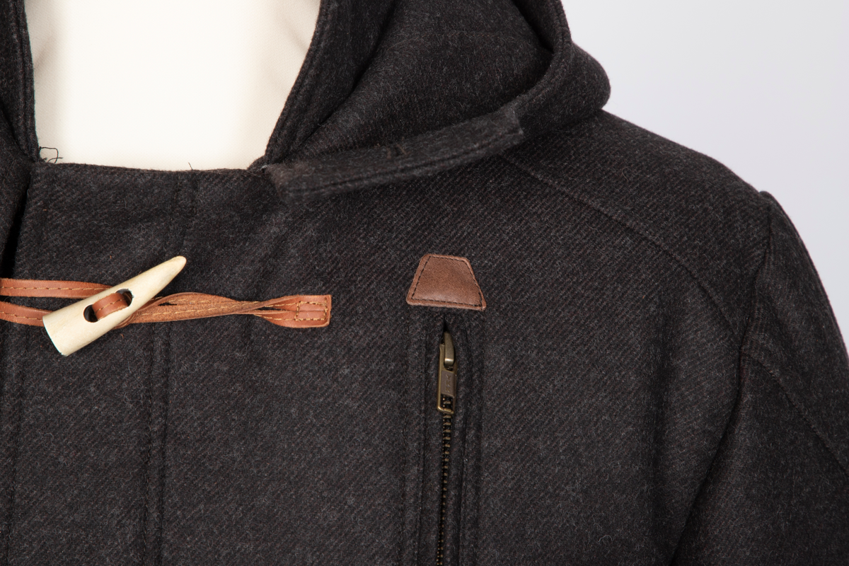 Dyffel med hette i brun ull. Bærestykke foran, på skuldrene og et bærestykke i skinnlignende materiale midt bak. Glidelås med klaff midt foran. Til lukking tre skinnsnorer med en beinlignende knapp. Store innfelte lommer med stor klaff med knapphull og gyllenbrun knapp. Søm midt bak med splitt, og formende panelsømmer. Belte festet rett foran sidesømmen i livet. Tosøms-ermer, dobbelklaff med knapp og knapphull og tre hemper på øverste klaffen. Sømmen på skulderens bærestykke fortsetter over til erme, parallelt nedover til den skrår inn tre centimeter før klaffene. Rødt, rutestukket, vattert fôr. Jakken er laget med nano-teknologi.