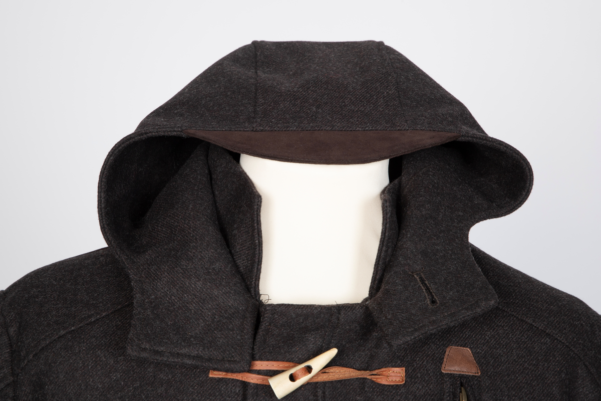 Dyffel med hette i brun ull. Bærestykke foran, på skuldrene og et bærestykke i skinnlignende materiale midt bak. Glidelås med klaff midt foran. Til lukking tre skinnsnorer med en beinlignende knapp. Store innfelte lommer med stor klaff med knapphull og gyllenbrun knapp. Søm midt bak med splitt, og formende panelsømmer. Belte festet rett foran sidesømmen i livet. Tosøms-ermer, dobbelklaff med knapp og knapphull og tre hemper på øverste klaffen. Sømmen på skulderens bærestykke fortsetter over til erme, parallelt nedover til den skrår inn tre centimeter før klaffene. Rødt, rutestukket, vattert fôr. Jakken er laget med nano-teknologi.