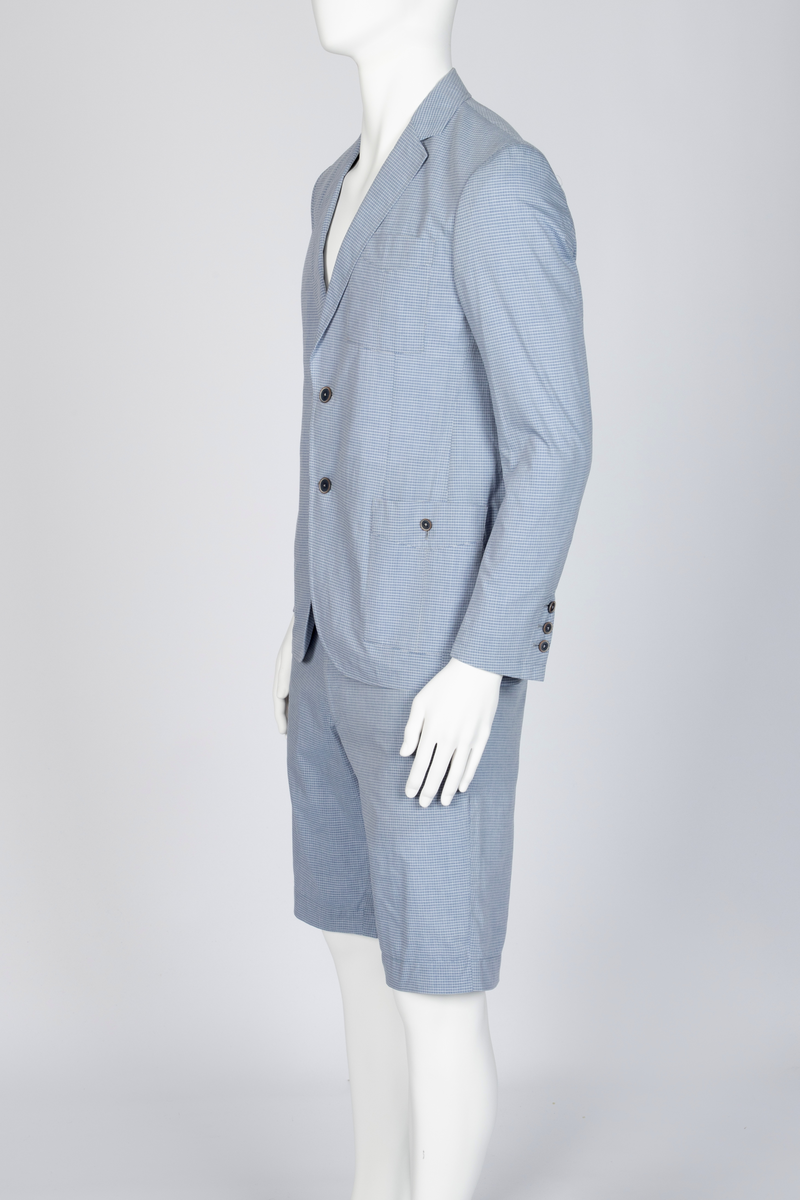 Herredress i smårutete, lyseblått og hvitt bomullsstoff bestående av kortbukse og jakke. Jakken er enkeltspent, med to knapper, har smale slag og krage og avrundede forbredder. Utenpåsatte lommer med nedstukket "klaff" i spiss med knapphull og knapp. Brystlomme. Ermene har åpen sliss med tre knapphull og knapper. Splitt midt bak. Fôr i ermer og nakken, samt to innvendige brystlommer lukket med knapp. Buksen er lårkort, har to stikklommer, en urlomme på venstre side og to baklommer med klaff. Linning med sju beltehemper, midt bak og over urlommen er det dobbel beltehempe. Gylf med glidelås og metallhektelukning i linningen.