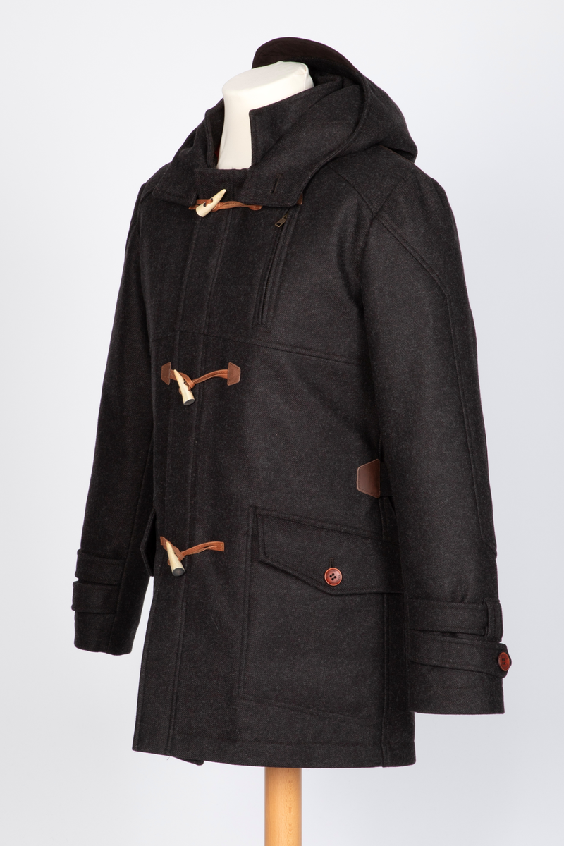 Dyffel med hette i brun ull. Bærestykke foran, på skuldrene og et bærestykke i skinnlignende materiale midt bak. Glidelås med klaff midt foran. Til lukking tre skinnsnorer med en beinlignende knapp. Store innfelte lommer med stor klaff med knapphull og gyllenbrun knapp. Søm midt bak med splitt, og formende panelsømmer. Belte festet rett foran sidesømmen i livet. Tosøms-ermer, dobbelklaff med knapp og knapphull og tre hemper på øverste klaffen. Sømmen på skulderens bærestykke fortsetter over til erme, parallelt nedover til den skrår inn tre centimeter før klaffene. Rødt, rutestukket, vattert fôr. Jakken er laget med nano-teknologi.