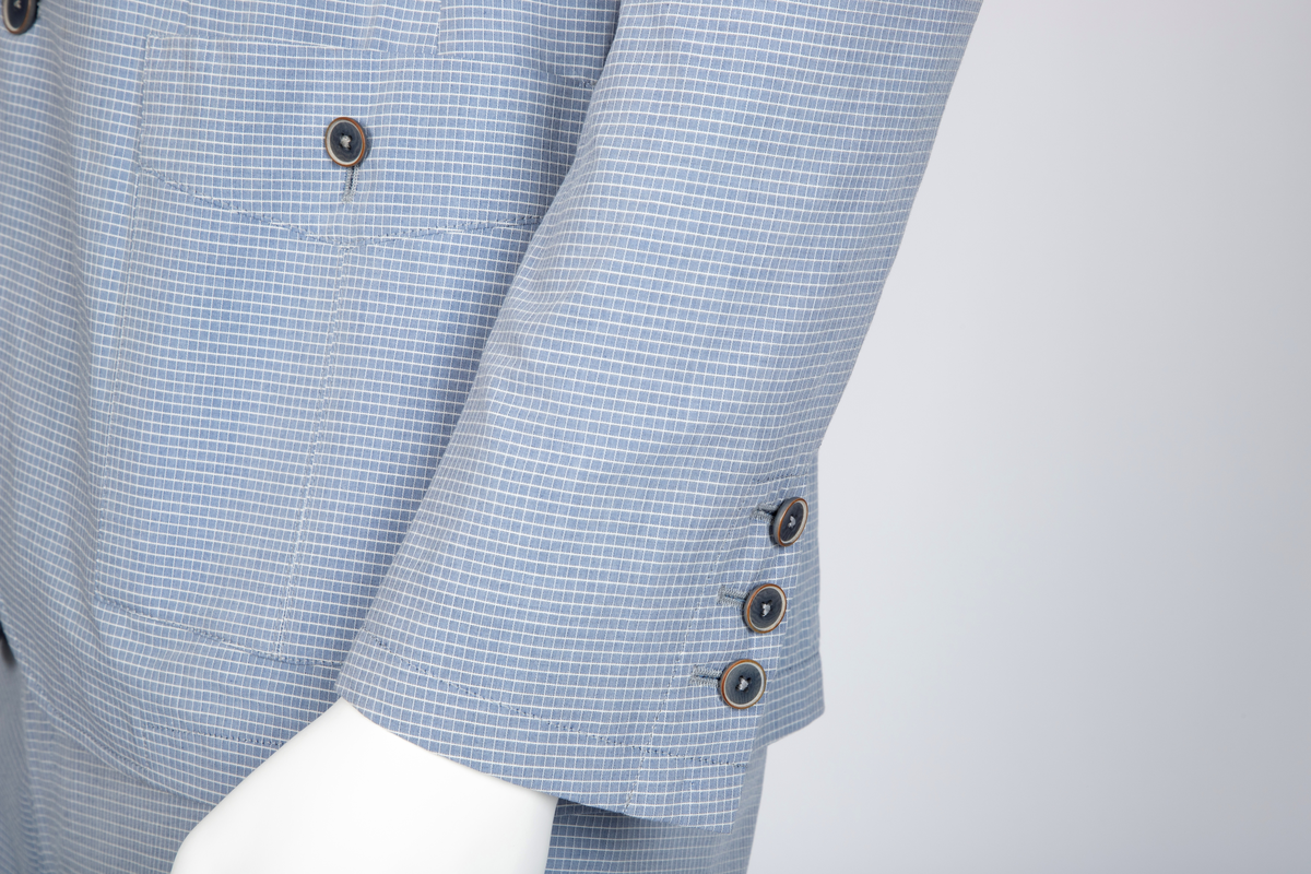 Herredress i smårutete, lyseblått og hvitt bomullsstoff bestående av kortbukse og jakke. Jakken er enkeltspent, med to knapper, har smale slag og krage og avrundede forbredder. Utenpåsatte lommer med nedstukket "klaff" i spiss med knapphull og knapp. Brystlomme. Ermene har åpen sliss med tre knapphull og knapper. Splitt midt bak. Fôr i ermer og nakken, samt to innvendige brystlommer lukket med knapp. Buksen er lårkort, har to stikklommer, en urlomme på venstre side og to baklommer med klaff. Linning med sju beltehemper, midt bak og over urlommen er det dobbel beltehempe. Gylf med glidelås og metallhektelukning i linningen.