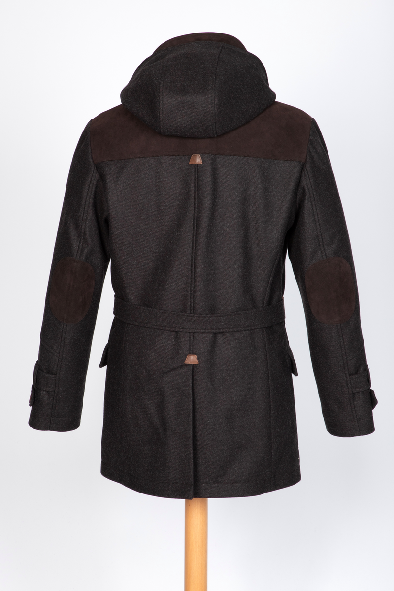 Dyffel med hette i brun ull. Bærestykke foran, på skuldrene og et bærestykke i skinnlignende materiale midt bak. Glidelås med klaff midt foran. Til lukking tre skinnsnorer med en beinlignende knapp. Store innfelte lommer med stor klaff med knapphull og gyllenbrun knapp. Søm midt bak med splitt, og formende panelsømmer. Belte festet rett foran sidesømmen i livet. Tosøms-ermer, dobbelklaff med knapp og knapphull og tre hemper på øverste klaffen. Sømmen på skulderens bærestykke fortsetter over til erme, parallelt nedover til den skrår inn tre centimeter før klaffene. Rødt, rutestukket, vattert fôr. Jakken er laget med nano-teknologi.