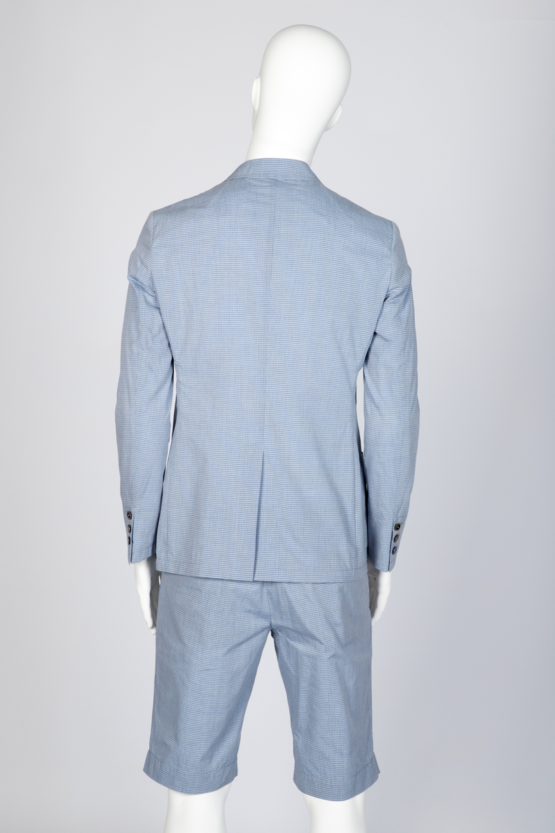 Herredress i smårutete, lyseblått og hvitt bomullsstoff bestående av kortbukse og jakke. Jakken er enkeltspent, med to knapper, har smale slag og krage og avrundede forbredder. Utenpåsatte lommer med nedstukket "klaff" i spiss med knapphull og knapp. Brystlomme. Ermene har åpen sliss med tre knapphull og knapper. Splitt midt bak. Fôr i ermer og nakken, samt to innvendige brystlommer lukket med knapp. Buksen er lårkort, har to stikklommer, en urlomme på venstre side og to baklommer med klaff. Linning med sju beltehemper, midt bak og over urlommen er det dobbel beltehempe. Gylf med glidelås og metallhektelukning i linningen.