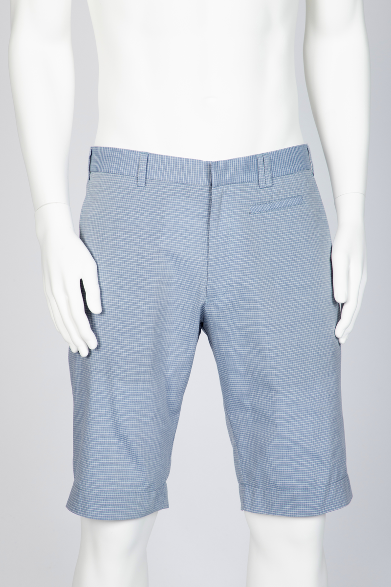 Herredress i smårutete, lyseblått og hvitt bomullsstoff bestående av kortbukse og jakke. Jakken er enkeltspent, med to knapper, har smale slag og krage og avrundede forbredder. Utenpåsatte lommer med nedstukket "klaff" i spiss med knapphull og knapp. Brystlomme. Ermene har åpen sliss med tre knapphull og knapper. Splitt midt bak. Fôr i ermer og nakken, samt to innvendige brystlommer lukket med knapp. Buksen er lårkort, har to stikklommer, en urlomme på venstre side og to baklommer med klaff. Linning med sju beltehemper, midt bak og over urlommen er det dobbel beltehempe. Gylf med glidelås og metallhektelukning i linningen.