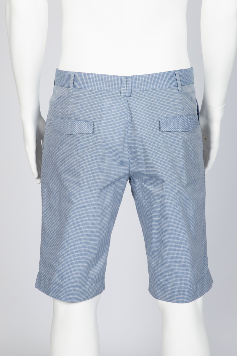 Herredress i smårutete, lyseblått og hvitt bomullsstoff bestående av kortbukse og jakke. Jakken er enkeltspent, med to knapper, har smale slag og krage og avrundede forbredder. Utenpåsatte lommer med nedstukket "klaff" i spiss med knapphull og knapp. Brystlomme. Ermene har åpen sliss med tre knapphull og knapper. Splitt midt bak. Fôr i ermer og nakken, samt to innvendige brystlommer lukket med knapp. Buksen er lårkort, har to stikklommer, en urlomme på venstre side og to baklommer med klaff. Linning med sju beltehemper, midt bak og over urlommen er det dobbel beltehempe. Gylf med glidelås og metallhektelukning i linningen.