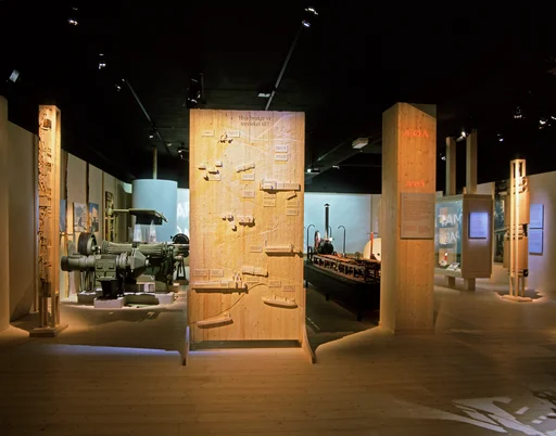 Fra rommet «Over hele verden» i Norsk skogmuseums basisutstilling «Tid for Skog», som ble bygd vinteren 2000-2001. Fotografiet viser en treskjerm – 110 centimeter bred og 220 centimeter høy – med tittelen «Hva bruker vi trevirket til?» Skjermen er utformet av Bo Jonzon, som var designer for den nevnte utstillingen. Øverst har han lagd en liten figur av en hogstmaskin, og ei liuta finérplate der årlig avvirkning i norske skoger angis i tall. Så følger små plater med informasjon om hvor store kvanta som eksporteres og importeres av rundtømmer. Deretter kan vi følge en tømmerbil som slynger seg nedover ei veglinje på skjermen. På vegen får vi vite hvor mye av tømmeret som årlig går til vedfyring, hvor mye som går til trelastindustrien (inkludert import- og eksporttall, samt oppgave over salg av flis). Deretter er det en montasje om sponplatefabrikkenes virkesforbruk og treforedlingsindustriens virkesforbruk og eksportkvanta. Nederst på skjermen kommer lastebilen til et havneområde med lasteskip. Ettersom markedet for tømmer og produkter fra den skogbaserte industrien er i kontinuerlig endring, har det vært et problem for museet å justere tallinformasjonen ofte nok.