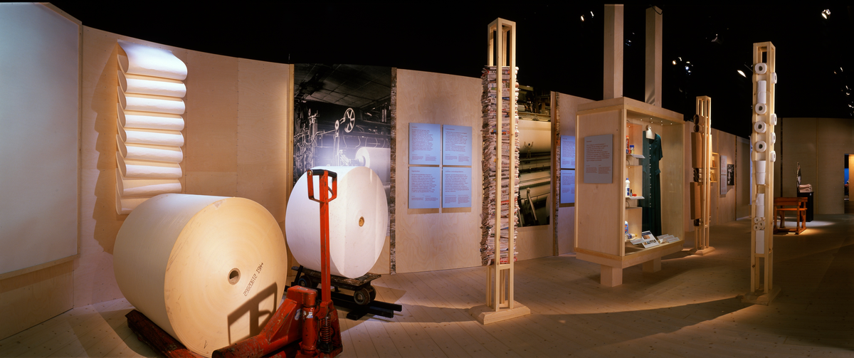 Fra rommet «Over hele verden» i Norsk skogmuseums basisutstilling «Tid for Skog», som ble bygd vinteren 2000-2001. Dette fotografiet viser montasjen som introduserer treforedlingsindustriene. På veggen bak de to store papirrullene i forgrunnen finner vi tekster som introduserer ulike greiner innenfor denne industien:

«Tresliperier
Fra 1840-åra ble det lagd papir ved å omdanne trestokken til tremasse i en slipeprosess. Den våte slipemassen ble viderebehandlet og resulterte i et skjørt, men billig papir. Det skulle bli basis for verdens aviser i tida framover. Økt etterspørsel etter tremasse i Europa og prisfall på trelast etter 1873 kan være en forklaring til vekst i tremasseproduksjonen. I 1870 sysselsatte tremassefabrikkene 61 arbeidere, i 1890 var tallet 2 045. Norge var i 1880-årene verdens ledende eksportør av tremasse, først og fremst til Storbritannia, der massen ble videreforedlet til papp og papir. Bentse Brug i Christiania startet landets første tresliperi i 1863.»

«Cellulosefabrikker
I 1880-åra ble en ny metode for å nyttiggjøre seg fibrene i tømmeret utviklet. I celluloseproduksjonen frigjøres fibrene i treet ved koking under trykk i kjemikalier. Cellulosen gav et meget høyverdig skrive- og fintrykkpapir fordi det kunne blekes. Aviser og bøker kunne ved hjelp av billig papir og forbedrete trykkemaskiner omsettes i større opplag enn tidligere. De fleste norske cellulosefabrikkene lå på Østlandet. I 1892 begynte Borregaard å produsere cellulose, og i 1895 stod de for drøyt 30 % av Norges Celluloseproduksjon.»

«Papirfabrikker
En del tremassefabrikker begynte å produsere cellulose. Våt tremasse var kostbar å frakte, og den kunne bare lagres i begrenset tid før den begynte å mugne. Cellulosen derimot kunne tørkes og lagres lenger. A/S Union i Skien startet som tremassefabrikk, men etter reorganisering av bedriften, startet byggingen av Europas største sulfittcellulosefabrikk i 1890. Borregaard startet papirproduksjon i 1896. I 1909 var det 28 papirfabrikker i Norge.»

«Arbeidere i treforedlingsindustrien
Sagbruks- og treforedlingsindustrien var Norges største industriarbeidsplass mellom 1860 og 1890. Mange steder i Norge, men særlig langs Drammenselva og i byene i Østfold ble treforedlingsindustri en stor arbeidsplass for tusener av arbeidere. I dag er treforedlingen omstrukturtert til færre bedrifter som produserer mer ved hjelp av datastyrte produksajonssystemer. Dette krever færre arbeidere, men til gjengjeld arbeidskraft med høy utdannelse.»

I glasskapet til høyre i bildet er det samlet en del eksempler på trekjemiske produkter fra den norske treforedlingsindustrien.