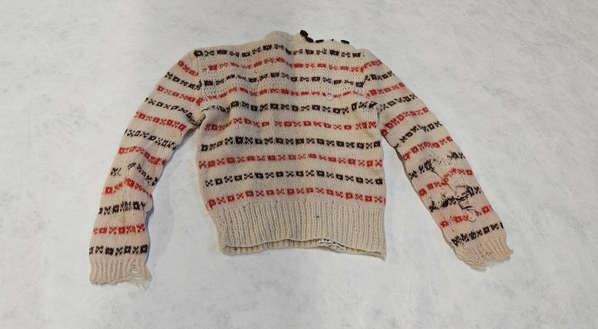 Genser med knepping på skulderen, fem brune blomsterknapper på høyre side. Mønster av firkanter og kryss.